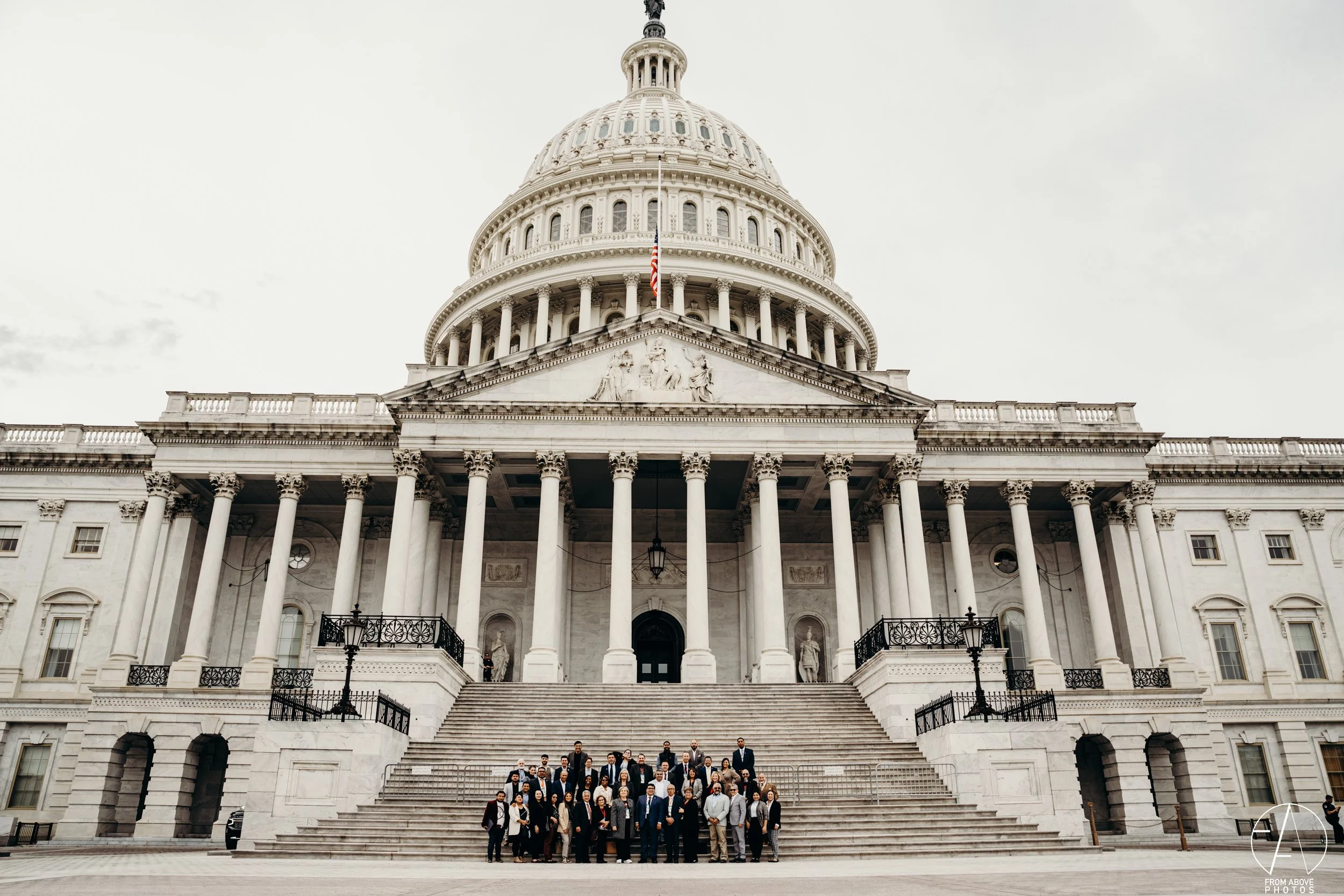 A large group of people standing on the steps of the United States Capitol building, which features a domed roof and neoclassical architecture, with an American flag flying in front.