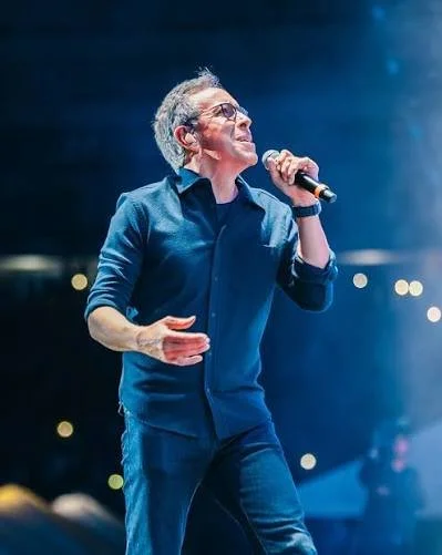 A man singing into a microphone on stage, wearing a denim jacket and in-ear monitors, with blue stage lighting in the background.
