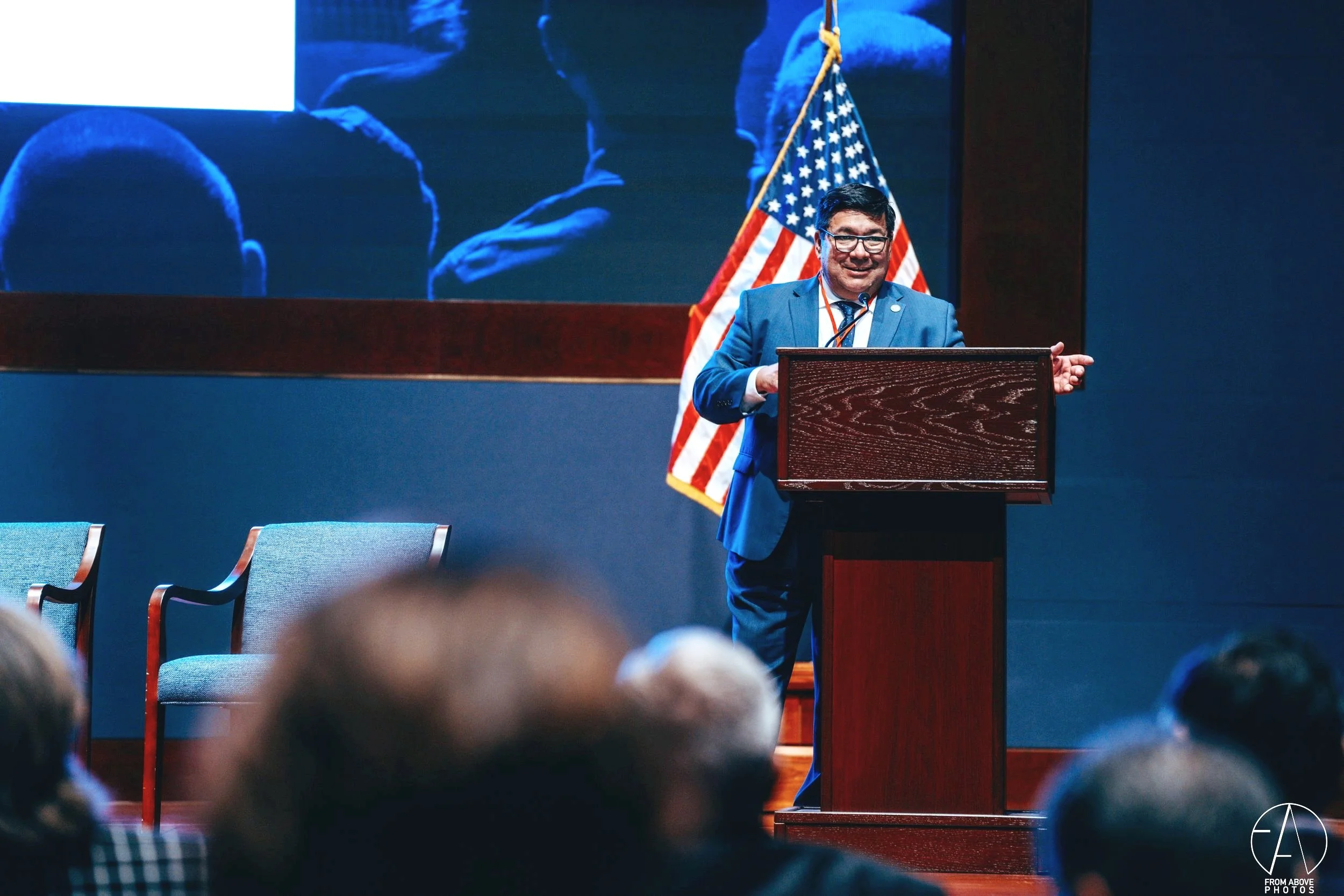 A man in a blue suit stands at a podium giving a speech with an American flag behind him. Audience members are seated in front of him.