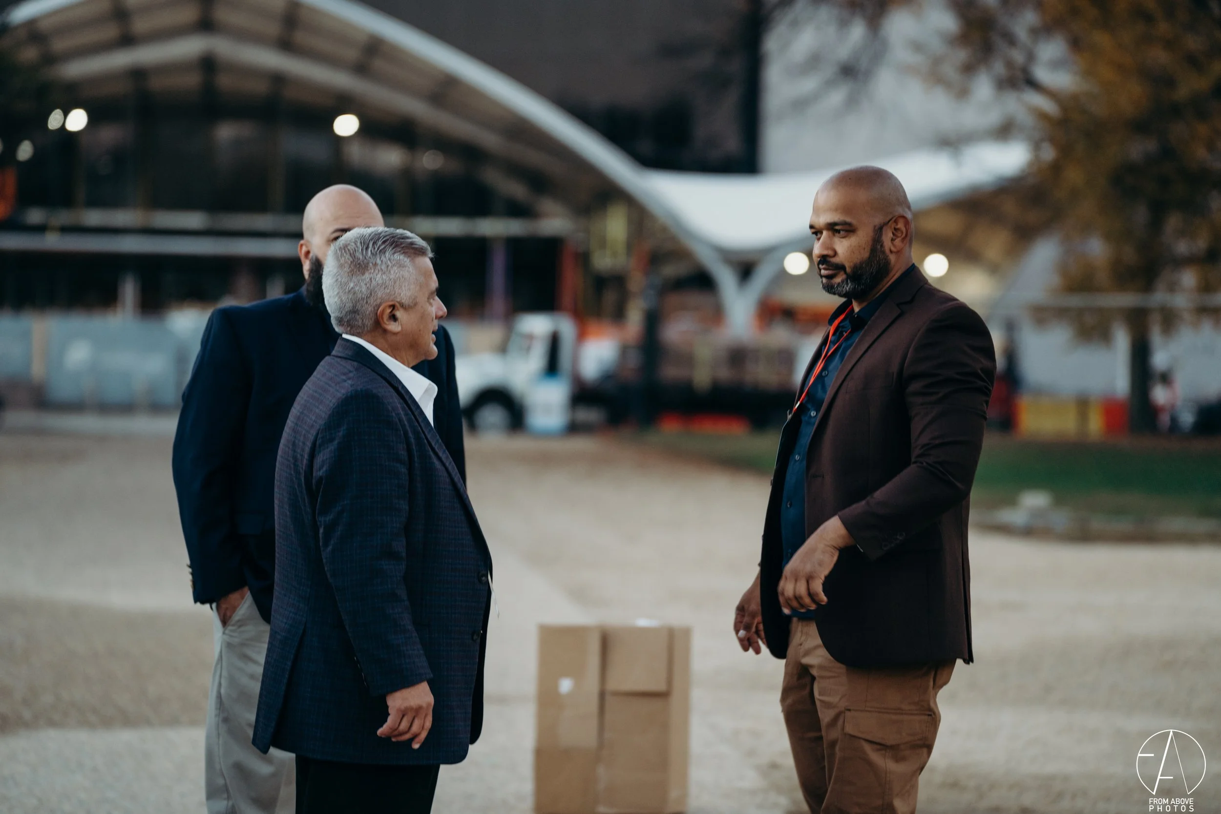 Three men in suits having a conversation outdoors during evening, with a modern building and parking lot in the background.