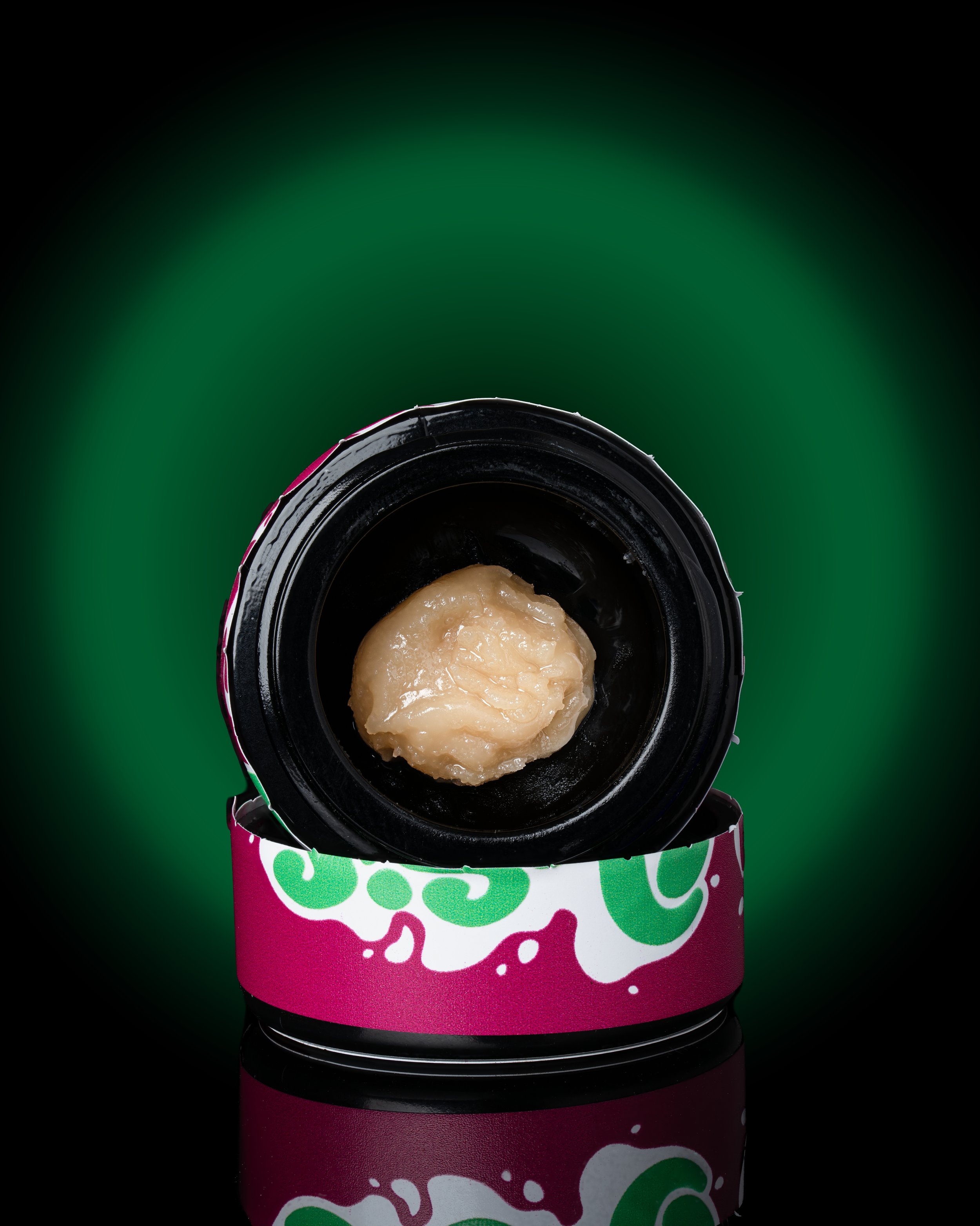 The image shows the top view of a black bottle with a black cap, containing a beige, viscous substance. The bottle has a colorful label with green and pink patterns, and is set against a dark green gradient background.
