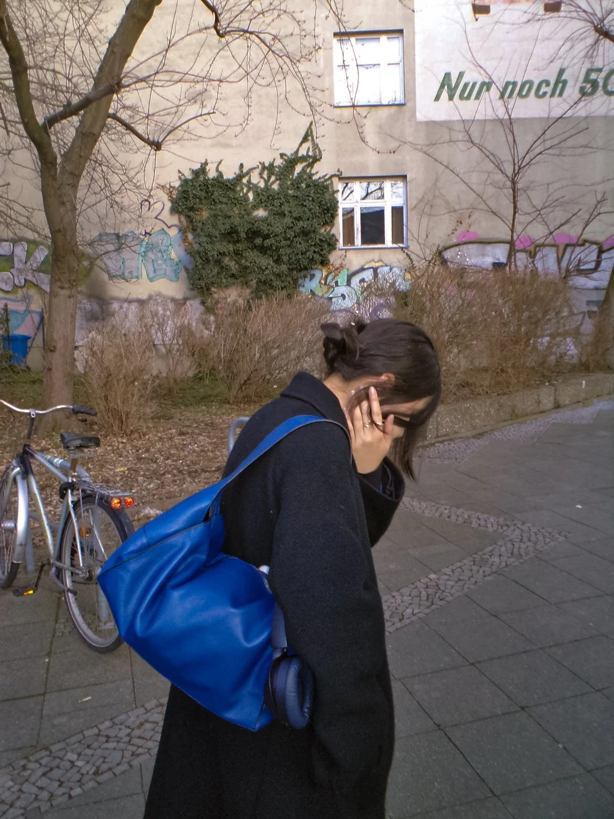 Woman with dark hair in a bun covering her face, wearing black coat and blue backpack, standing on a sidewalk in front of a building with graffiti, leafless trees, and a bicycle.