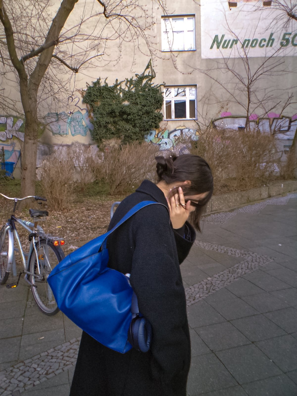 A woman with long dark hair, wearing a black coat and glasses, is standing on a sidewalk with her hand covering part of her face. She carries a large blue backpack and a pair of headphones dangle from her pocket. There is a bicycle parked behind her, and some leafless bushes and a tree are visible in the background. The wall behind has graffiti and a sign with German text.