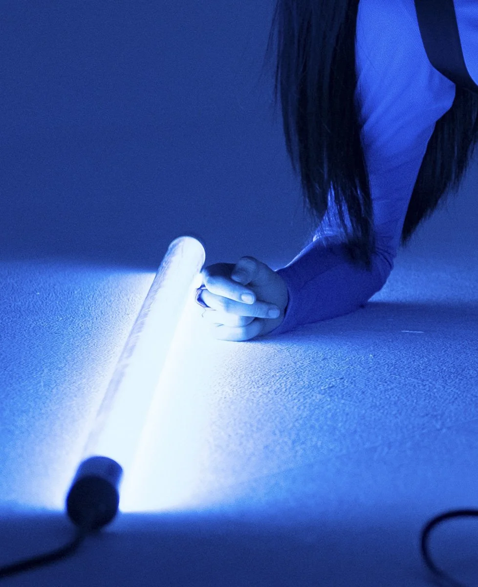 Person holding a flashlight or light tube on the floor, illuminating the surface with blue light, with long hair visible and wearing a long-sleeved shirt.
