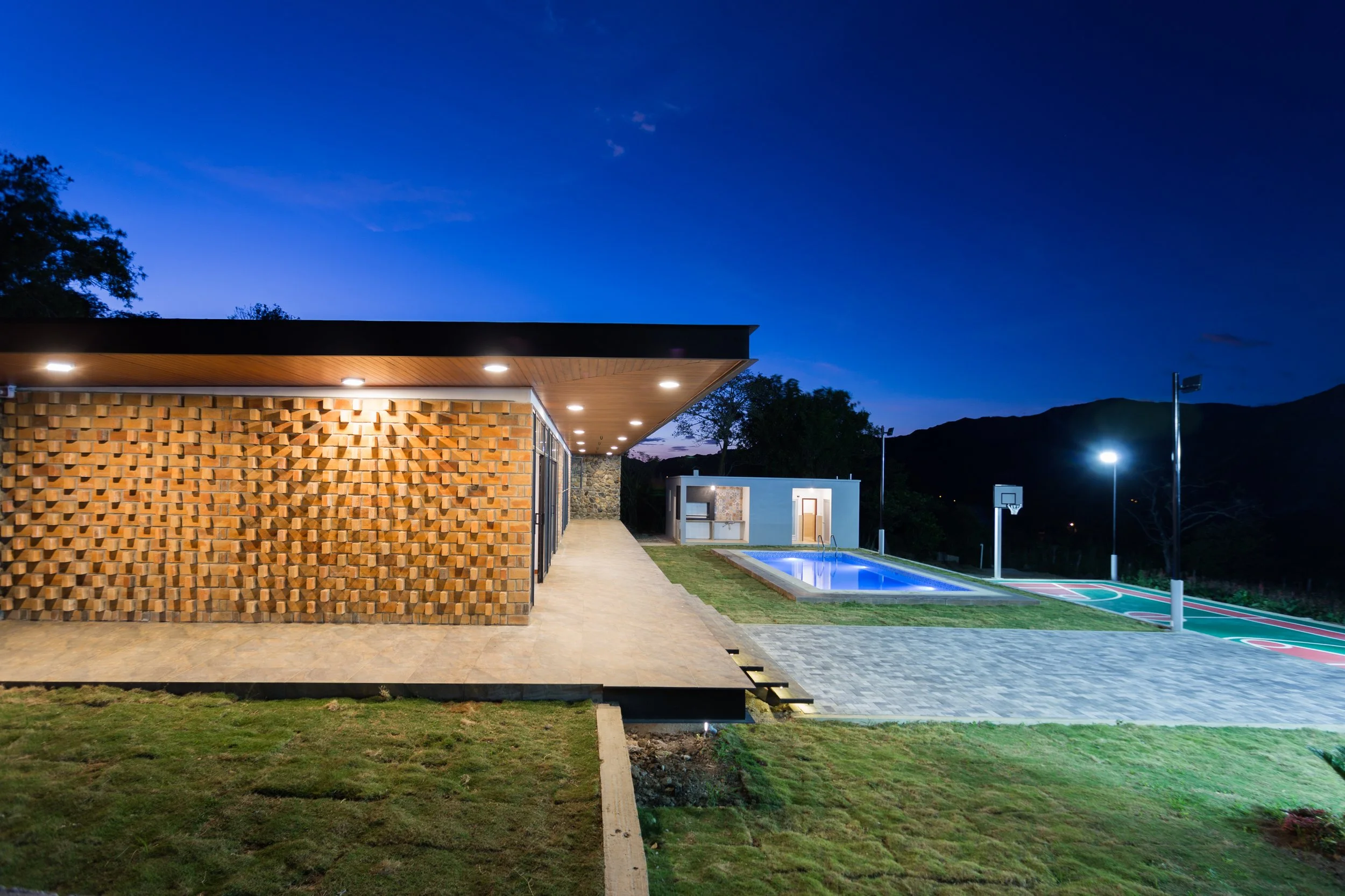 Casa moderna con piscina y cancha de baloncesto, iluminación nocturna, en un entorno natural con montañas al fondo.