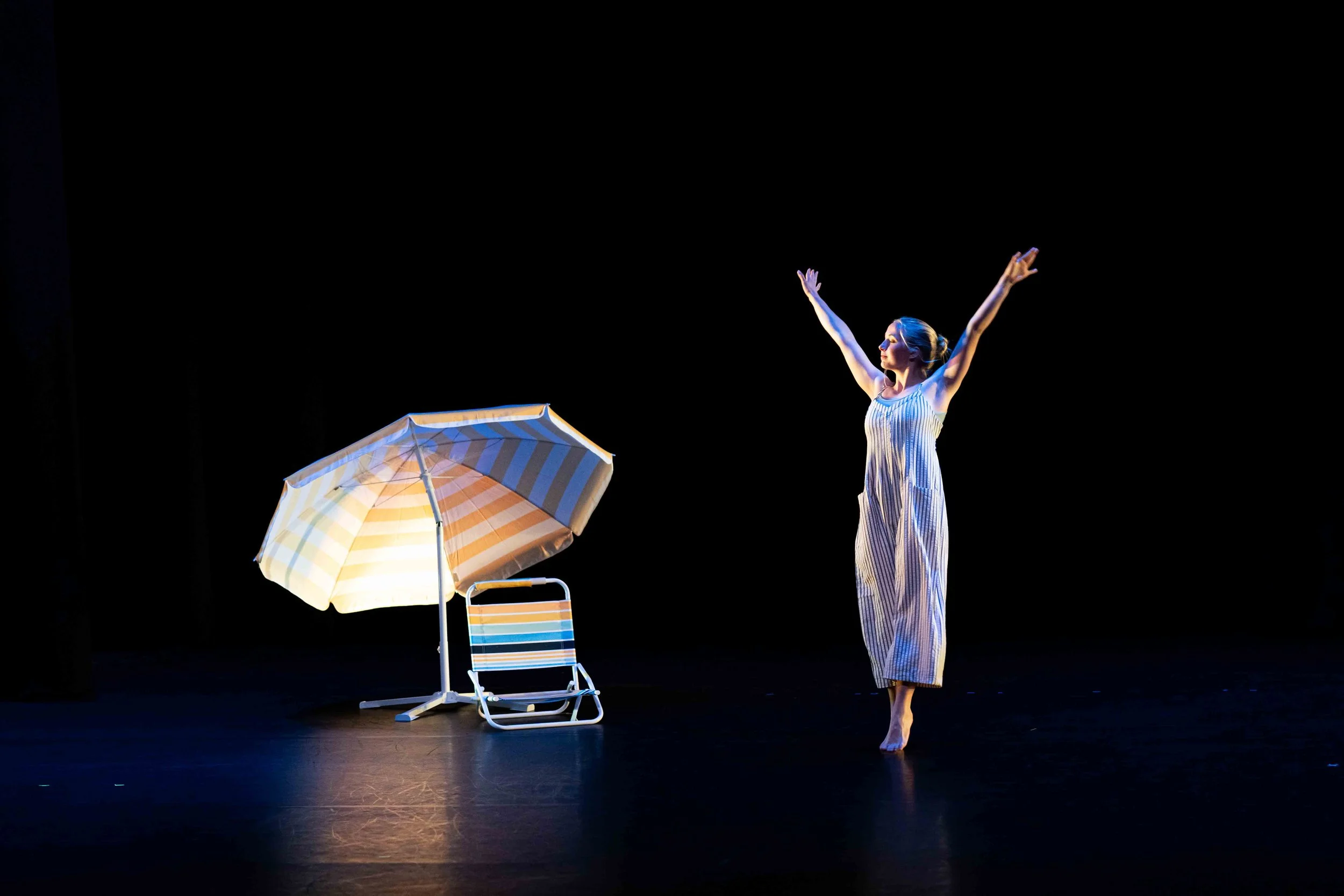 Dancer in releve with arms stretched above at high diagonals. A beach chair and umbrella are on the left side of the photo.