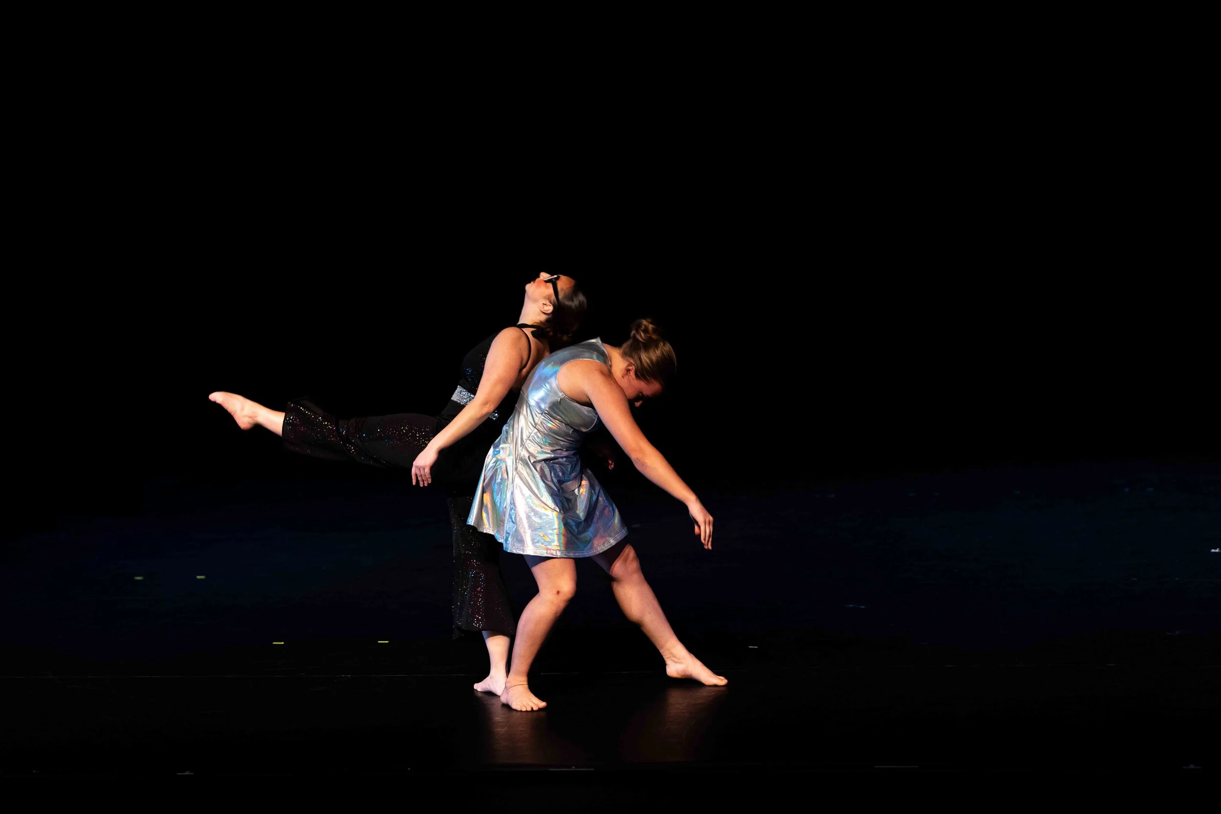 One dancer in a black jumpsuit is leaning back, supported by the back of another dancer in a silver dress.
