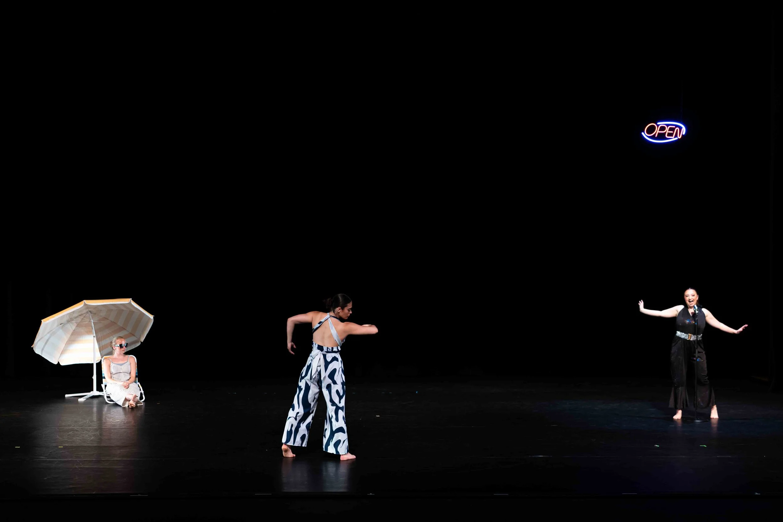 Three dancers across stage: the left one in sunglasses, sitting in a low beach chair; the center one facing upstage in a pose looking toward a bent right arm; the right one seemingly singing into a microphone, with a neon "open" sign above her head.
