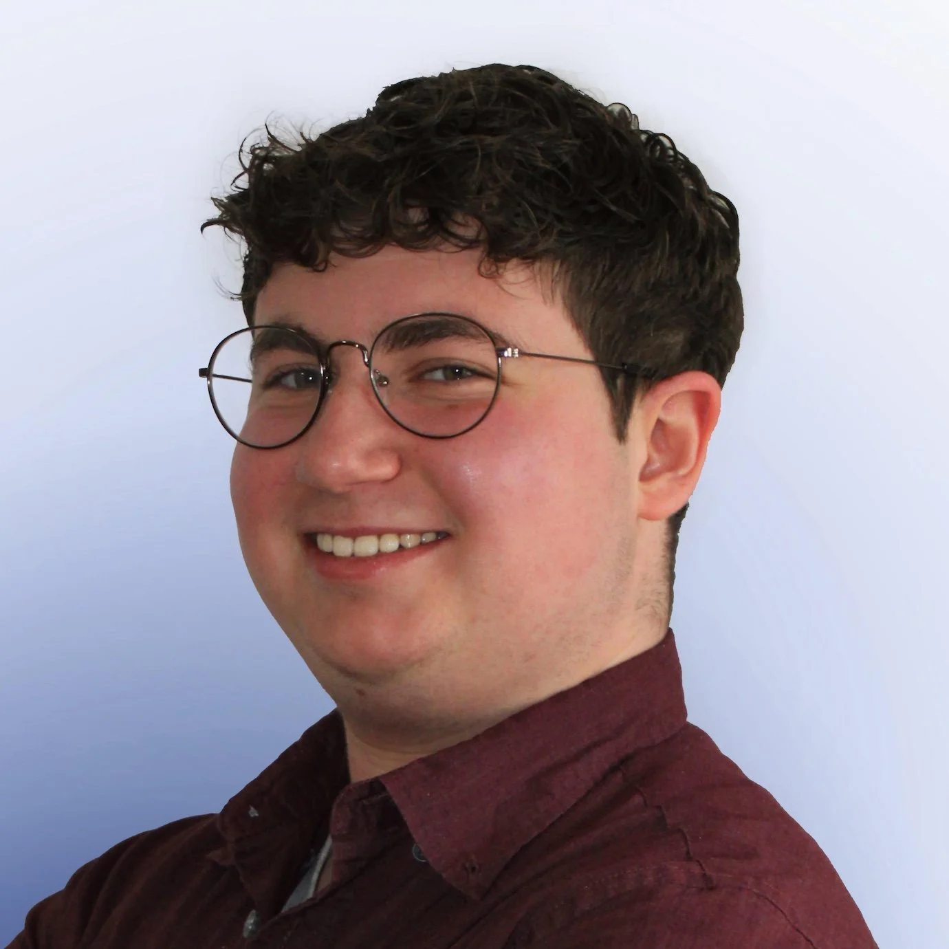 Young man with curly dark hair, glasses, and a friendly smile, wearing a maroon shirt, against a light blue background.