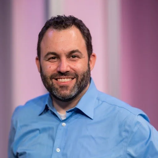 A smiling man with dark hair and a beard, wearing a light blue button-up shirt, standing in front of a pink and blue backdrop.