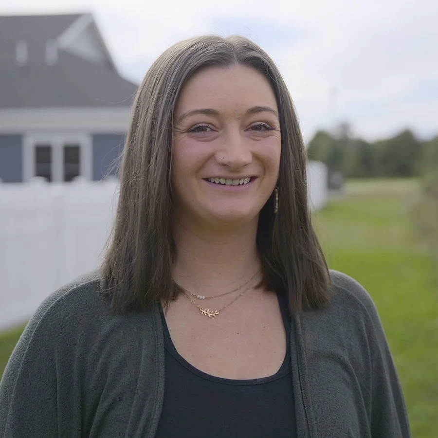 A woman with shoulder-length brown hair smiling outdoors in front of a house and a grassy area.
