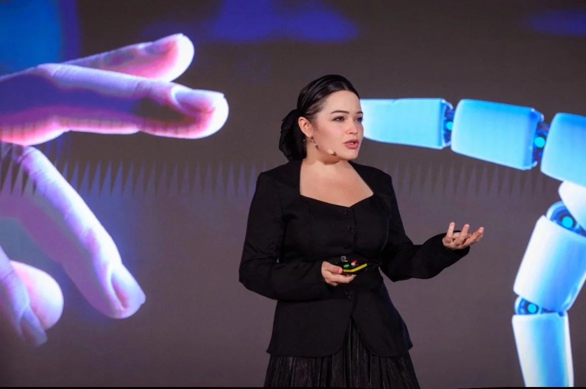 A woman with dark hair tied back, smiling slightly, sitting indoors in front of a neutral background with a plant partially visible on the left. Text overlays identify her as Emily Genatowski, a researcher in AI domestic robotics, in a televised interview.