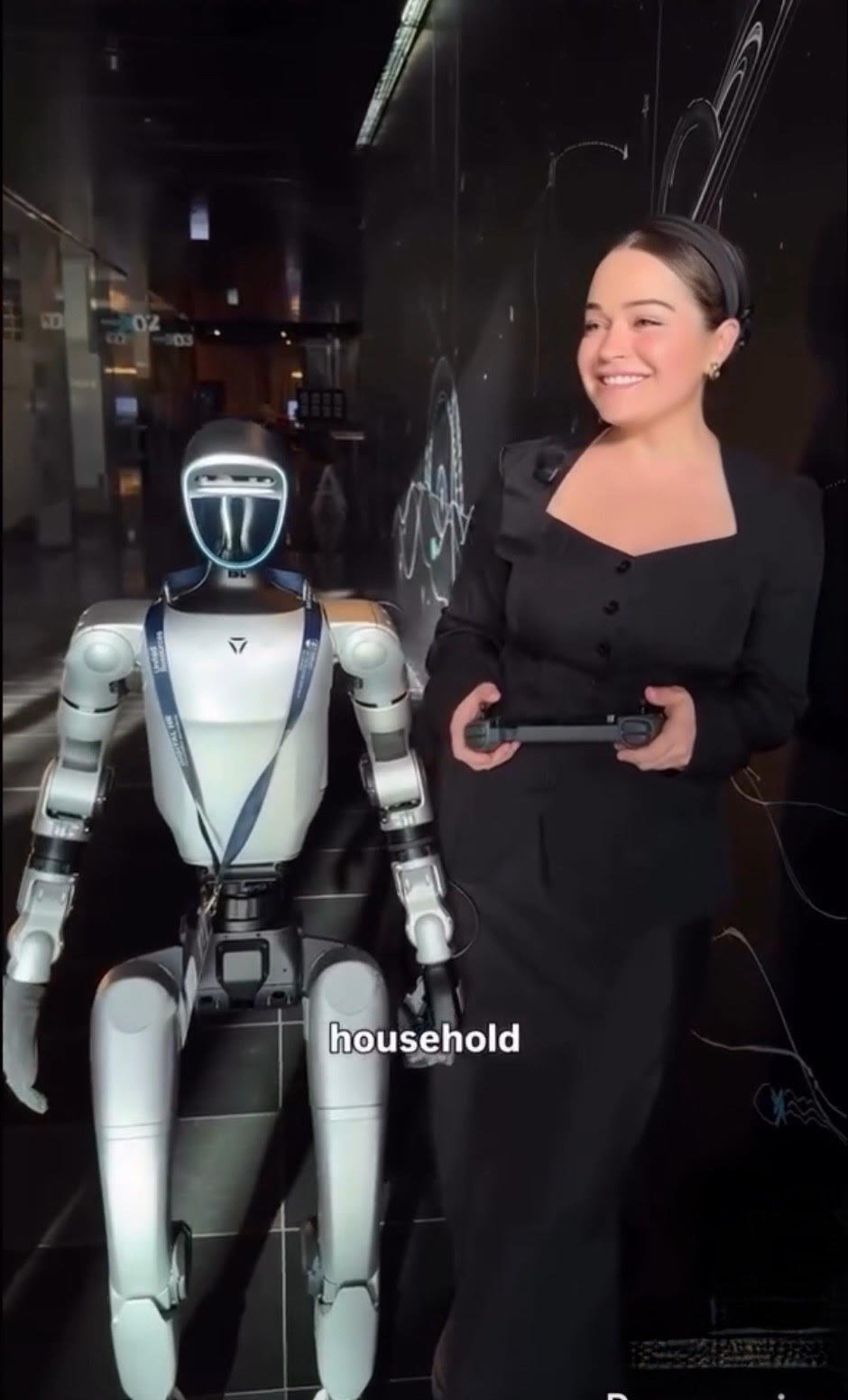 A woman with dark hair tied back, smiling slightly, sitting indoors in front of a neutral background with a plant partially visible on the left. Text overlays identify her as Emily Genatowski, a researcher in AI domestic robotics, in a televised interview.