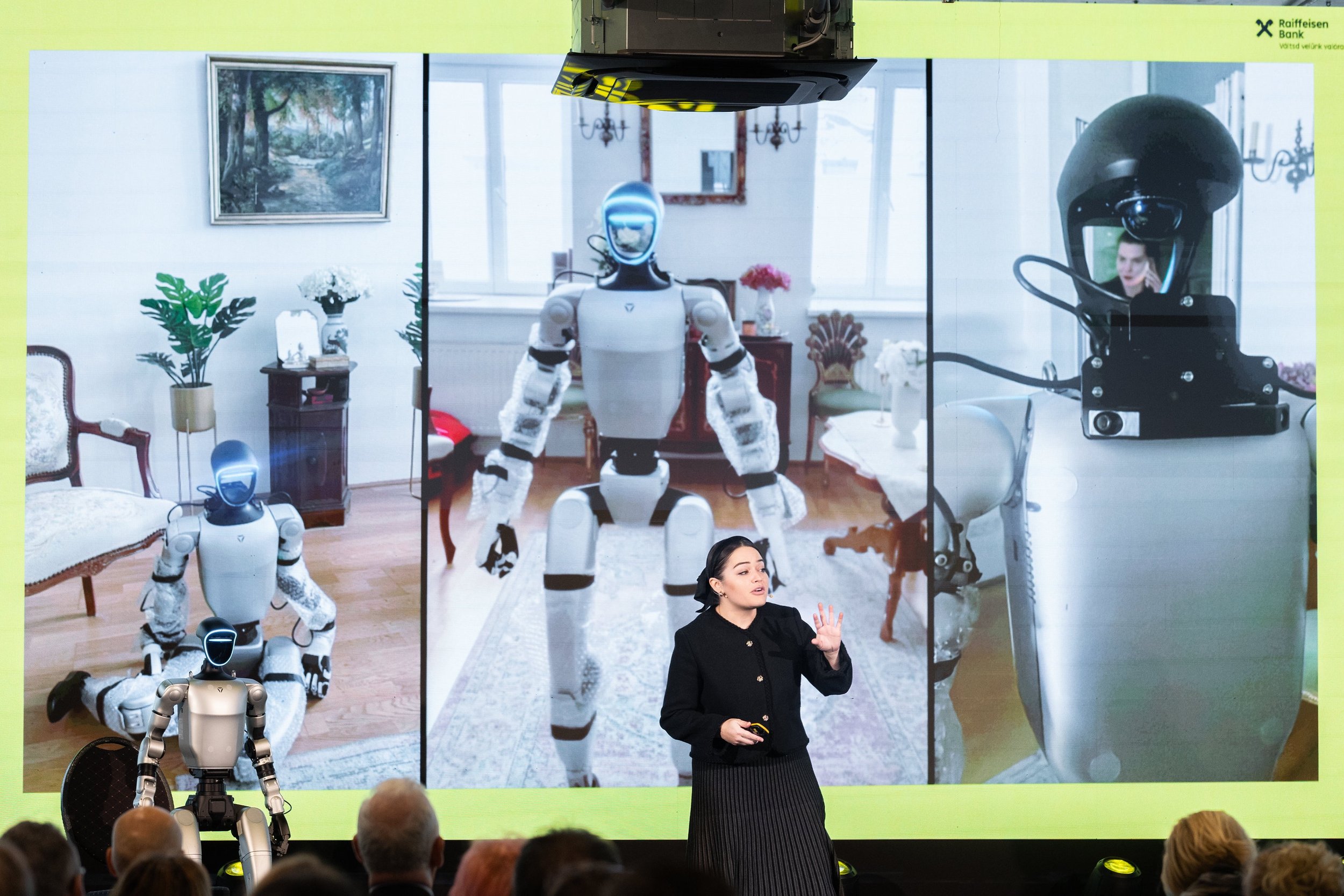 A woman on stage smiling and gesturing while standing next to a humanoid robot during a presentation or conference.