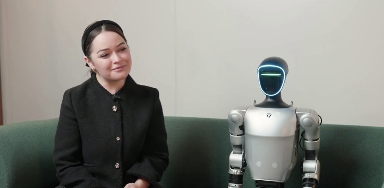A woman with dark hair tied back, smiling slightly, sitting indoors in front of a neutral background with a plant partially visible on the left. Text overlays identify her as Emily Genatowski, a researcher in AI domestic robotics, in a televised interview.