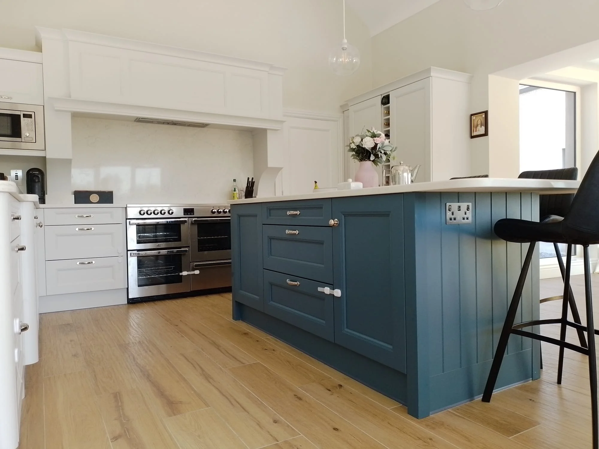 Modern kitchen with blue island, white cabinets, wood floor, double oven, barstools, and decorative vase on counter.