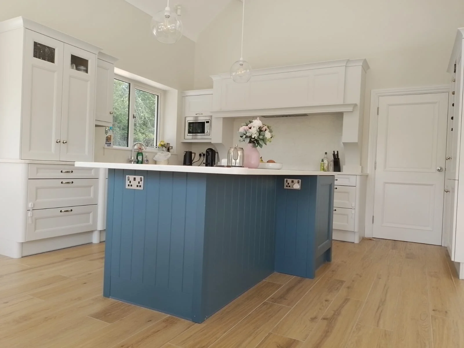 Modern kitchen with blue island and white cabinets, wooden floor, pendant lights, and stainless steel appliances.