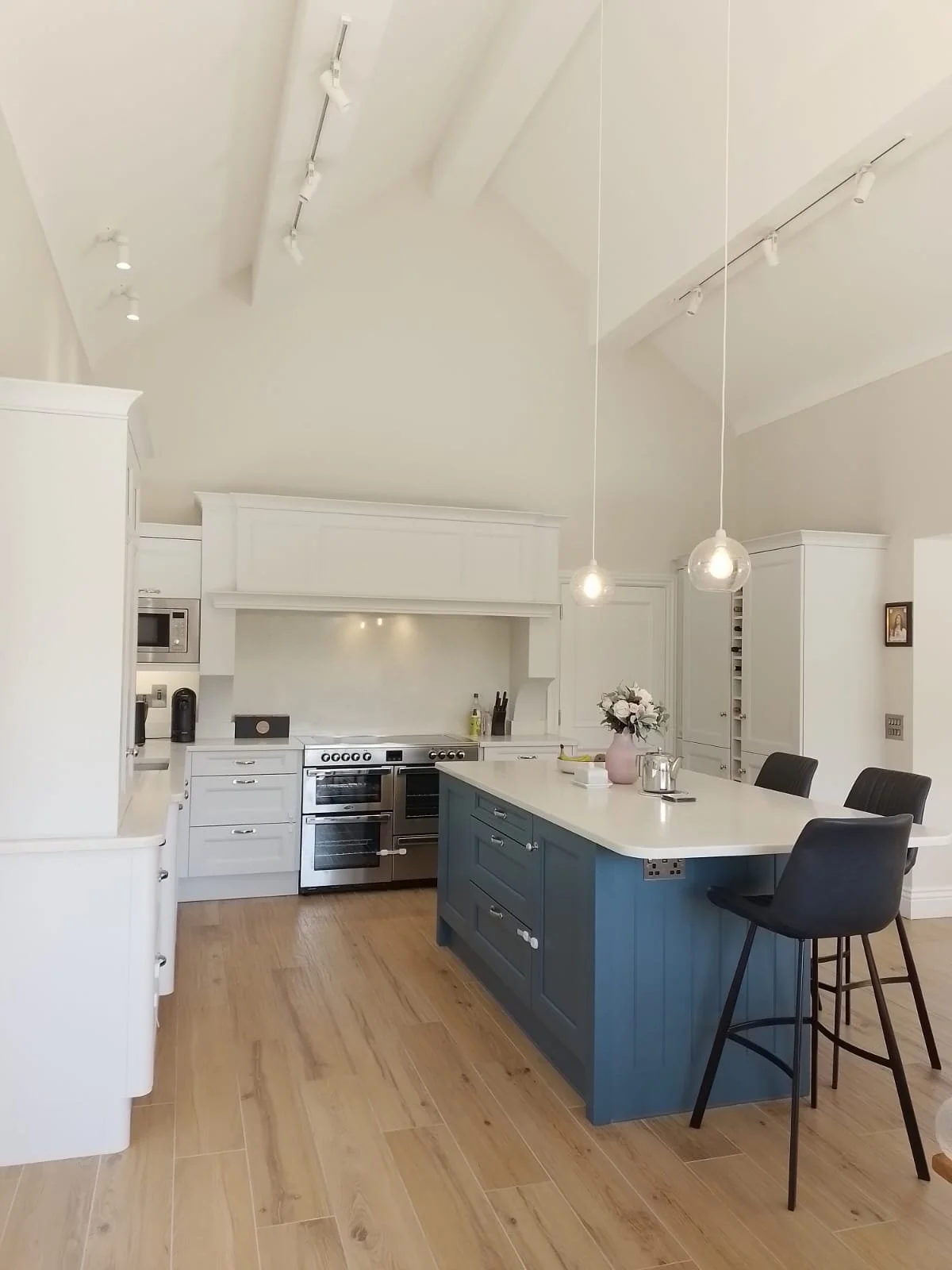 Modern kitchen interior with a large blue island, white cabinets, stainless steel oven, pendant lights, wooden flooring, and bar stools.