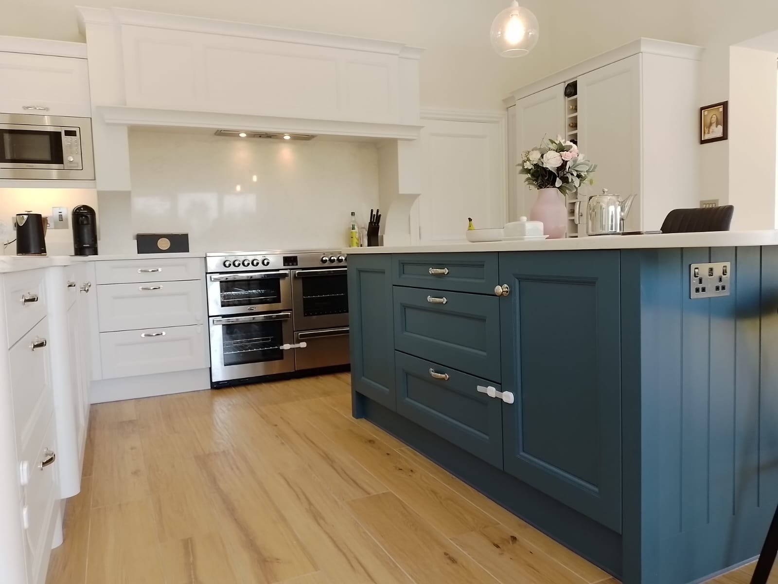 Modern kitchen with white cabinets, stainless steel oven and microwave, blue island with drawers, wooden floor, and overhead lighting. A teapot and flowers are on the island.