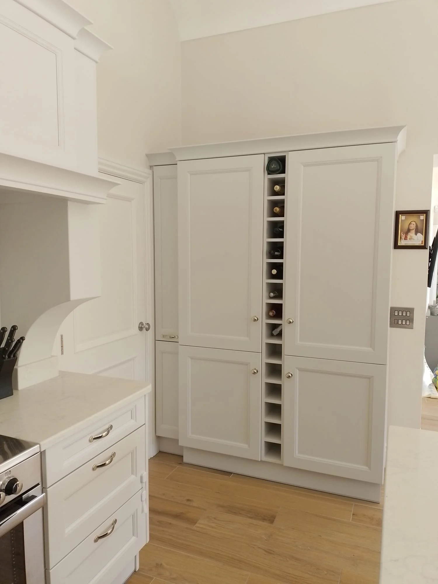 A modern kitchen corner with white cabinets, wooden floors, and a built-in wine rack. A countertop with drawers is visible, along with a knife block. There's a small framed picture on the wall.