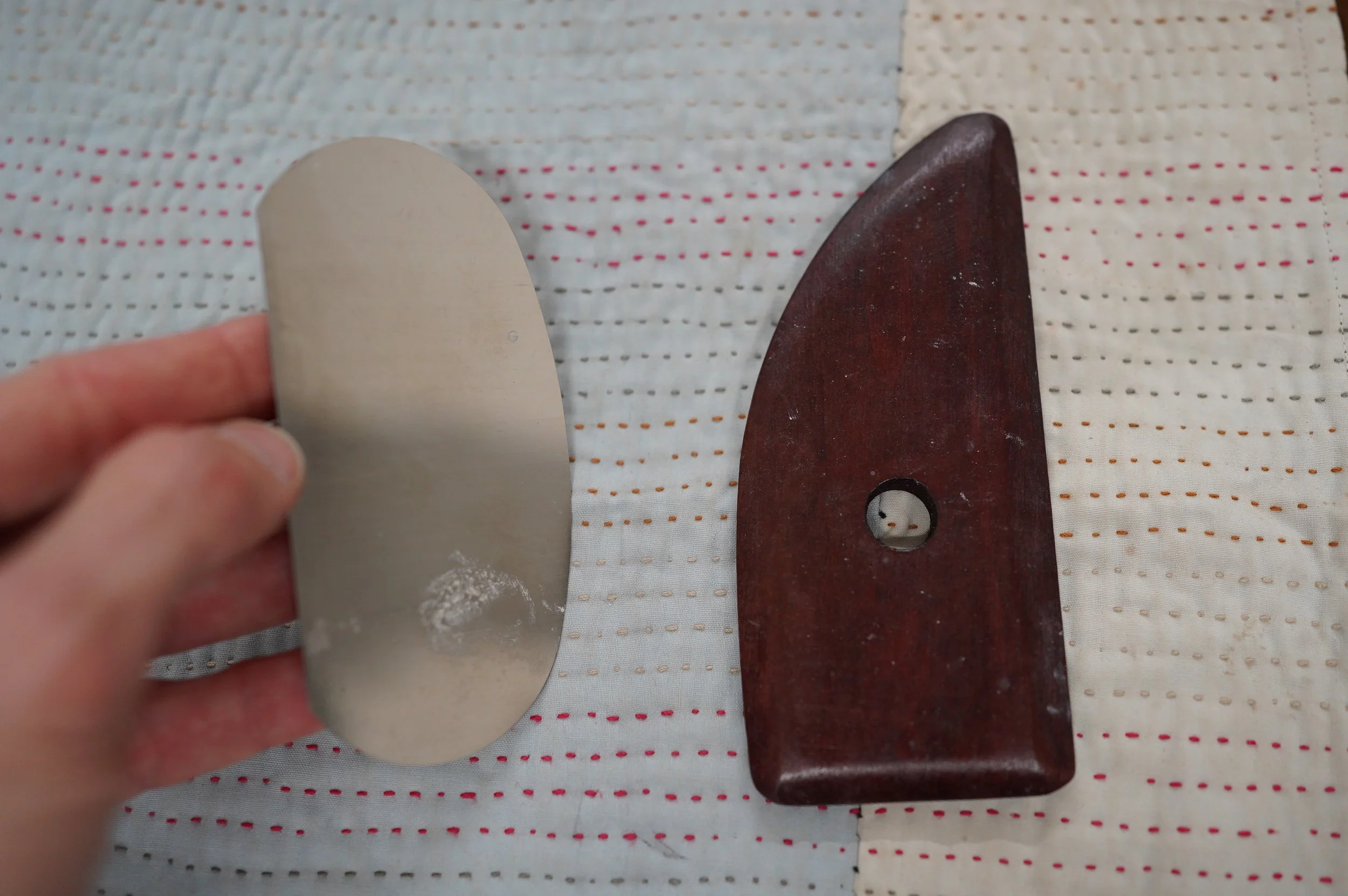A hand holds a metal rib tool for pottery next to a wooden rib tool.