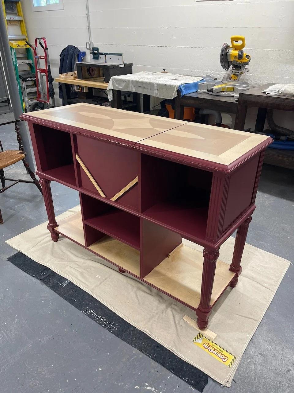 A wooden cabinet with a burgundy finish and decorative carved legs is in the process of being painted or finished, with some unpainted sections visible. The cabinet is placed on a beige fabric in a workshop with various tools and equipment in the background.
