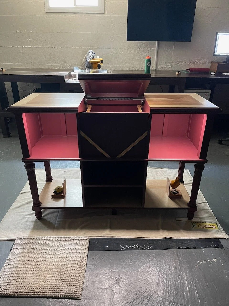 A partially assembled multi-compartment furniture piece with pink, black, and wood finishes, standing on a cloth on a concrete floor, with a small beige rug in front, in a workshop or garage setting.