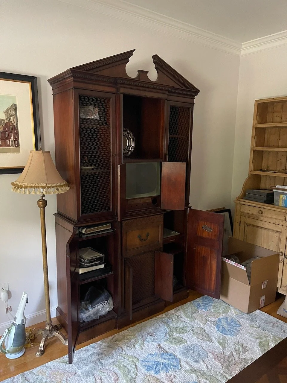 Wooden antique cabinet with open doors displaying shelves, a small TV inside, and various items stored inside and outside. There is a tall floor lamp to the left, a framed picture hanging on the wall, a cardboard box with items, a wooden bookshelf, and a patterned carpet on the floor.