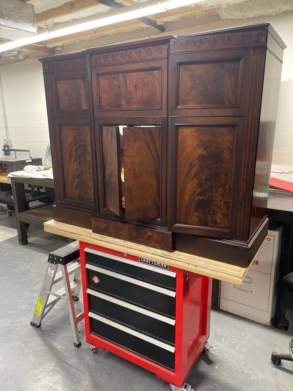 A wooden cabinet with decorative carvings on top, placed on a workbench in a workshop. The cabinet appears to be newly finished and is in the process of having doors installed.