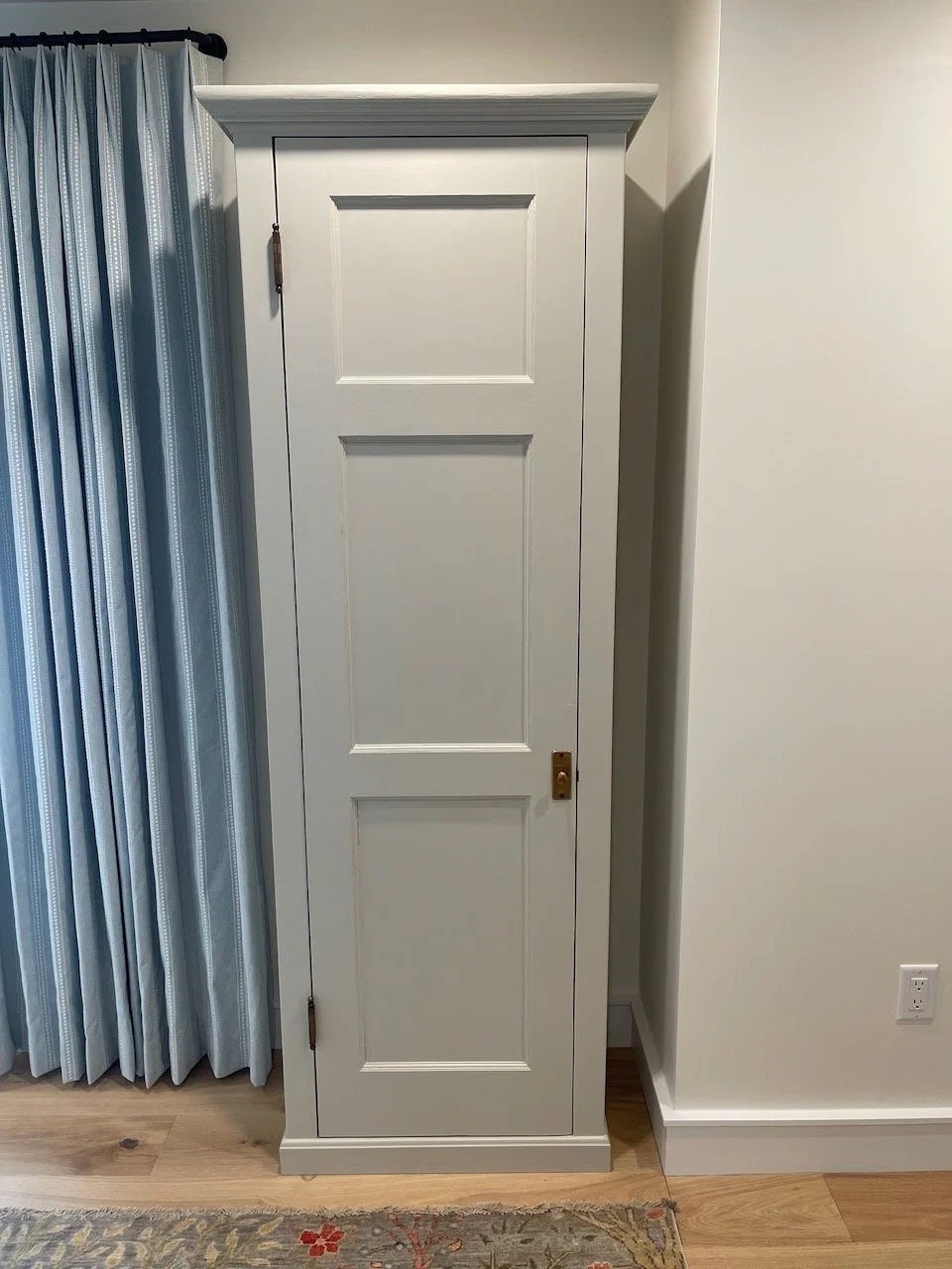 A white wooden cabinet with three recessed panels on the door, positioned next to a light-colored wall, with a curtain on the left side and a wooden floor beneath.