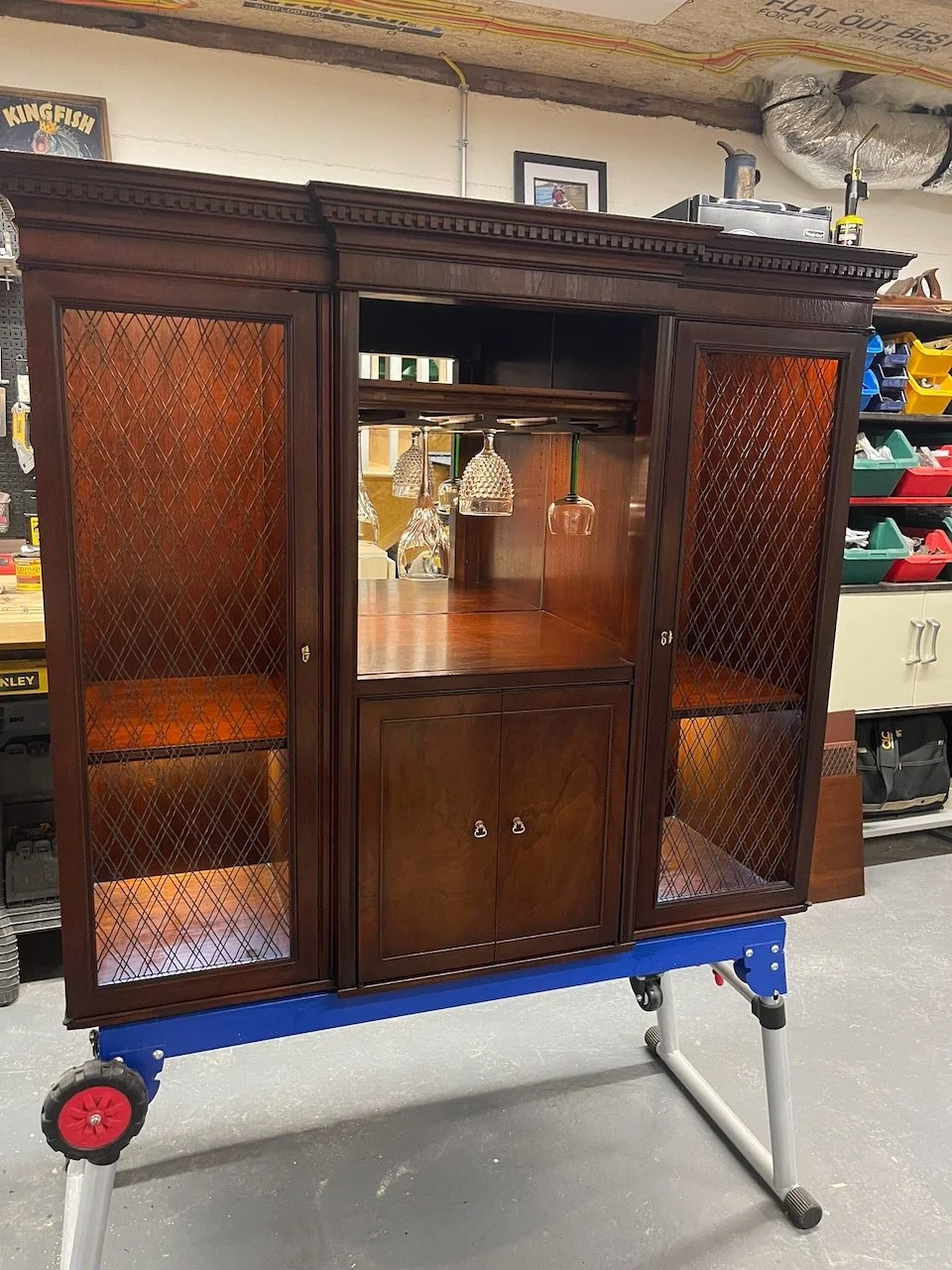 A vintage wooden bookshelf with glass front doors on a wheeled stand inside a store or workshop.