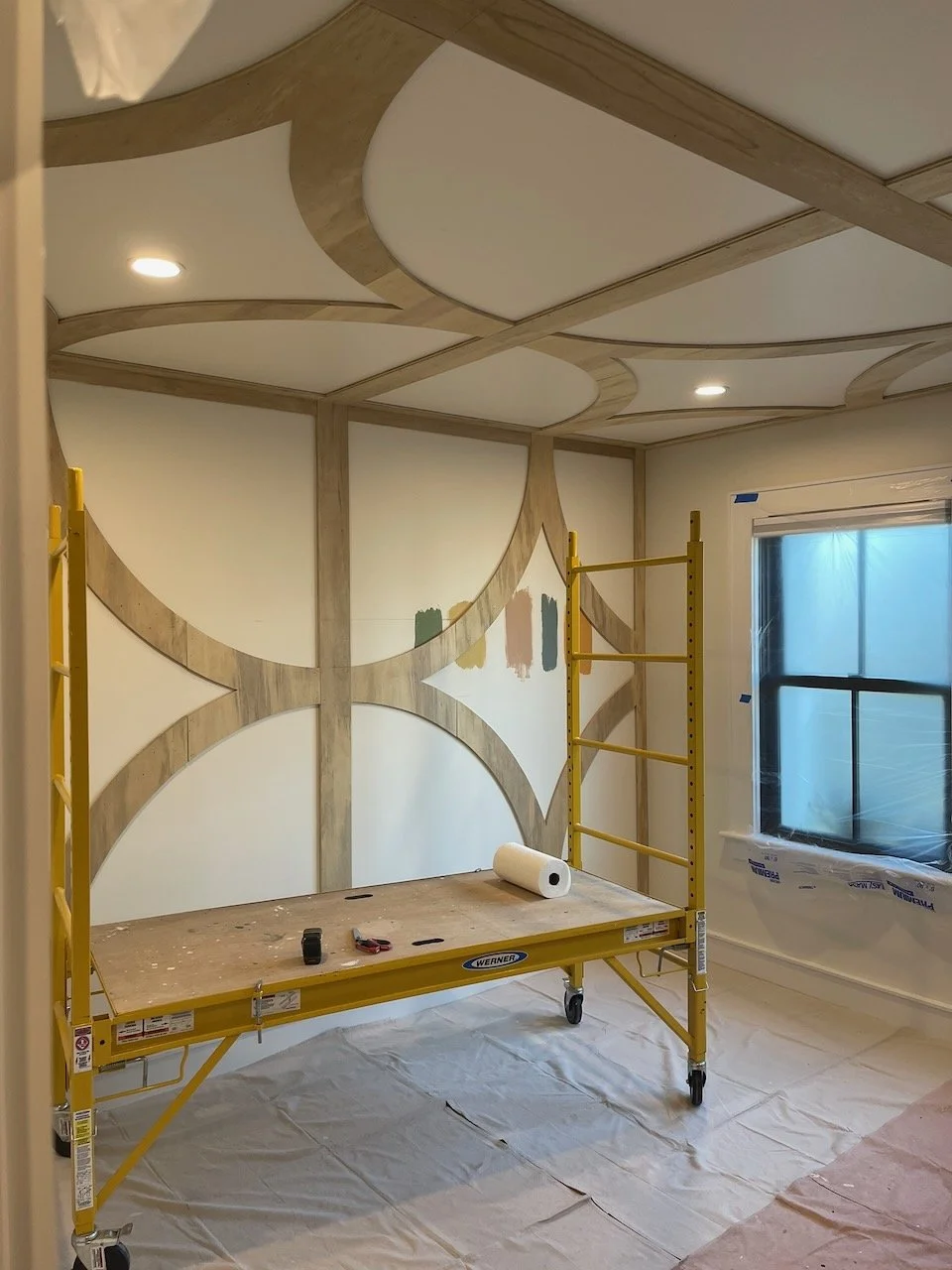 Room under construction with scaffolding, paint samples on the wall, and a window covered with plastic.