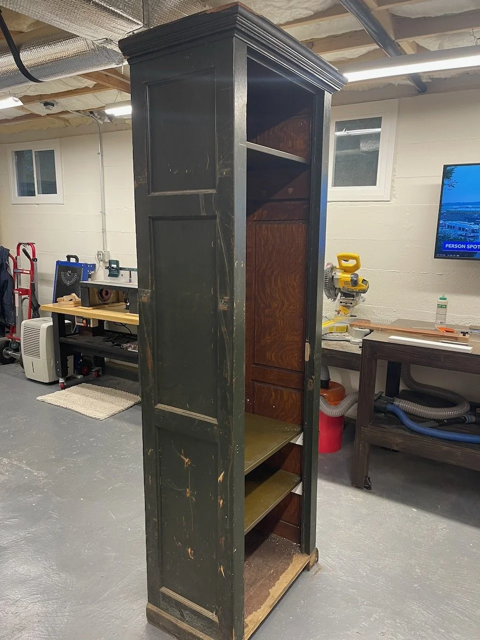 A tall, vintage wooden bookshelf with three shelves, painted black on the outside and with a dark brown interior, appears worn and scratched, situated in a workshop or basement.
