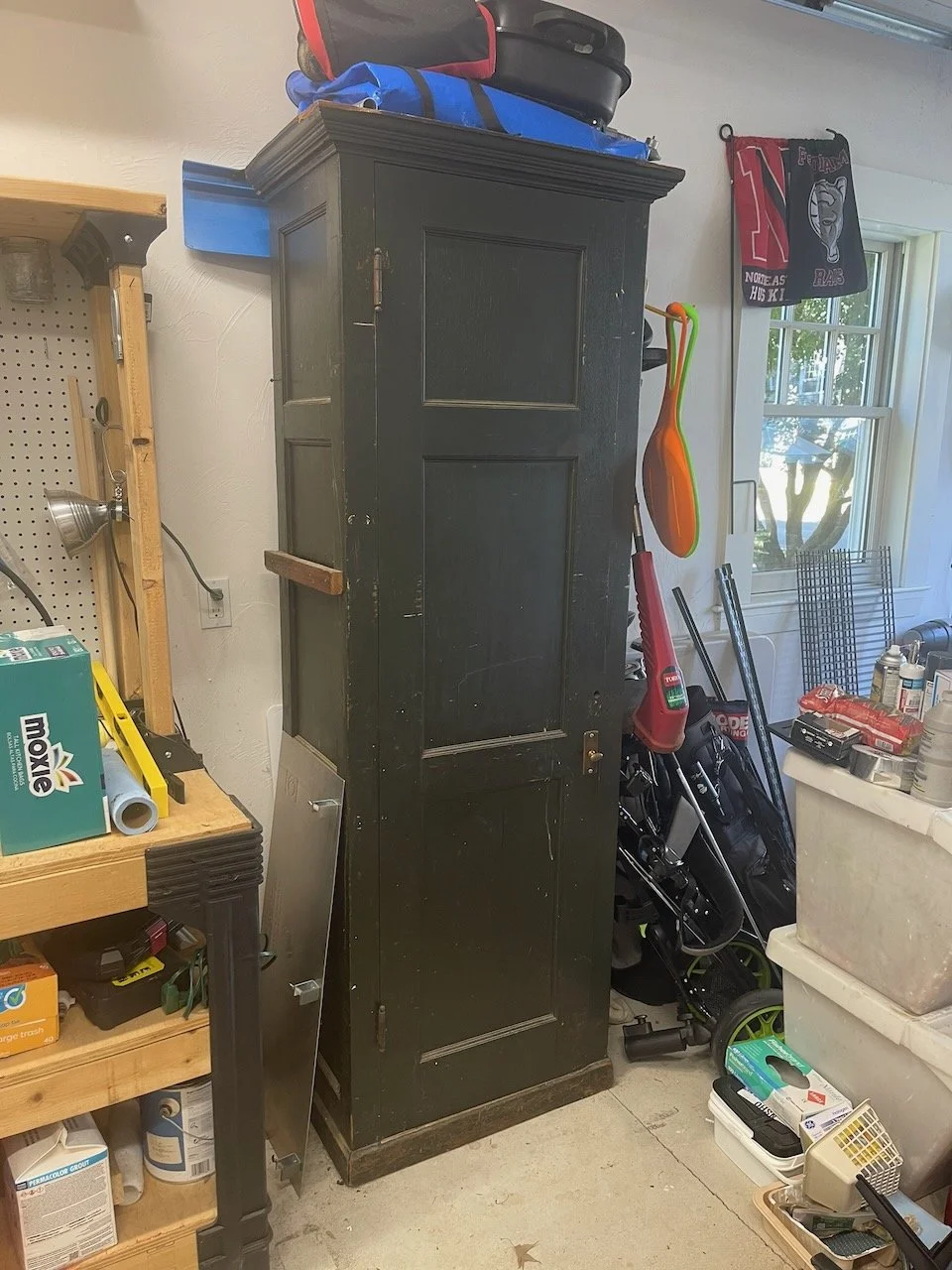 A tall black wooden cabinet in a cluttered garage with storage bins, tools, and sports equipment nearby.