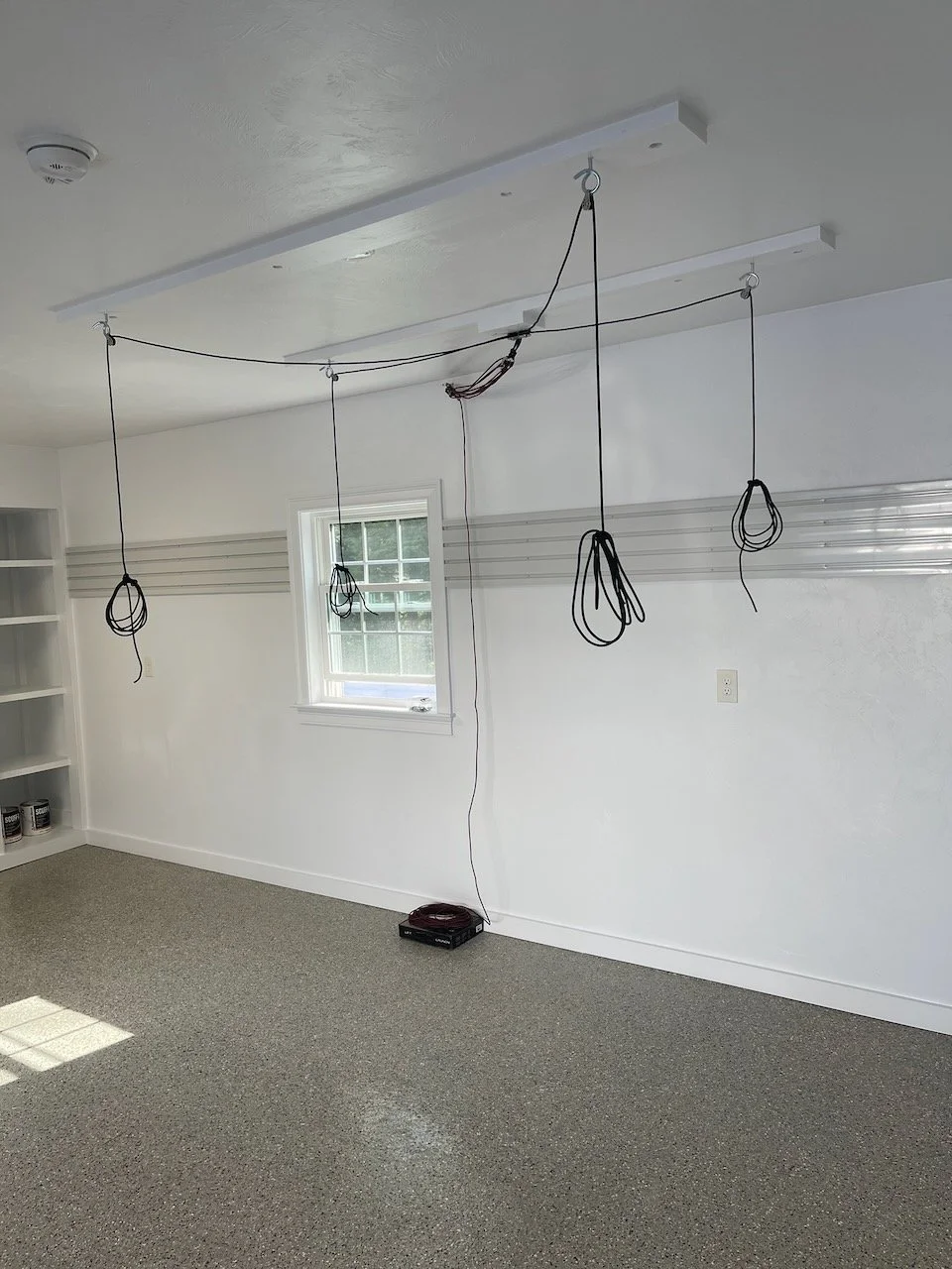 Room with wires hanging from ceiling, a window on the wall, shelf on the left, and a small black device on the floor.