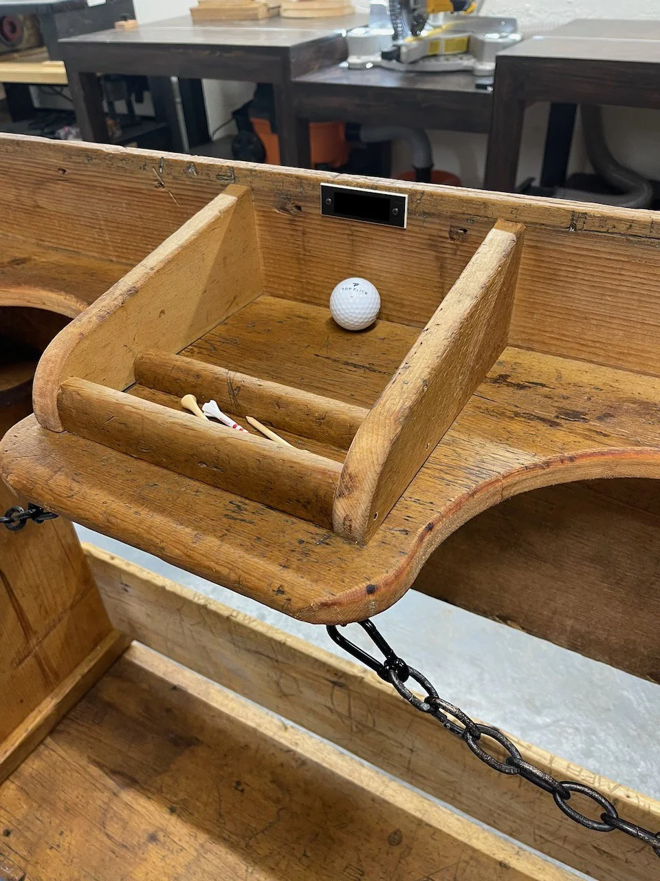A golf practice net with a golf ball and some golf tees inside, set on a wooden workbench in a workshop.