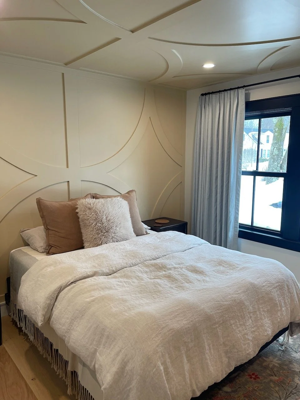A cozy bedroom featuring a bed with beige pillows, a fluffy white pillow, and a beige blanket. There is a window with white curtains and winter scenery outside.