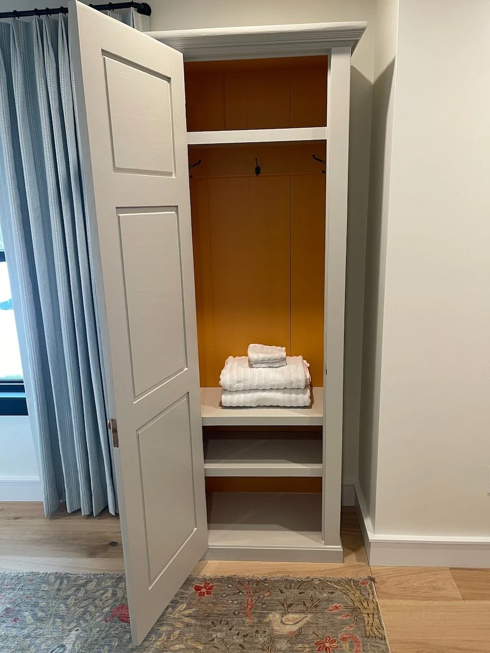 Empty white closet with a brown interior, two shelves, and a hanging rod. Folded white towels are stacked on the middle shelf. Outside the closet, light blue curtains and a window are partially visible on the left.