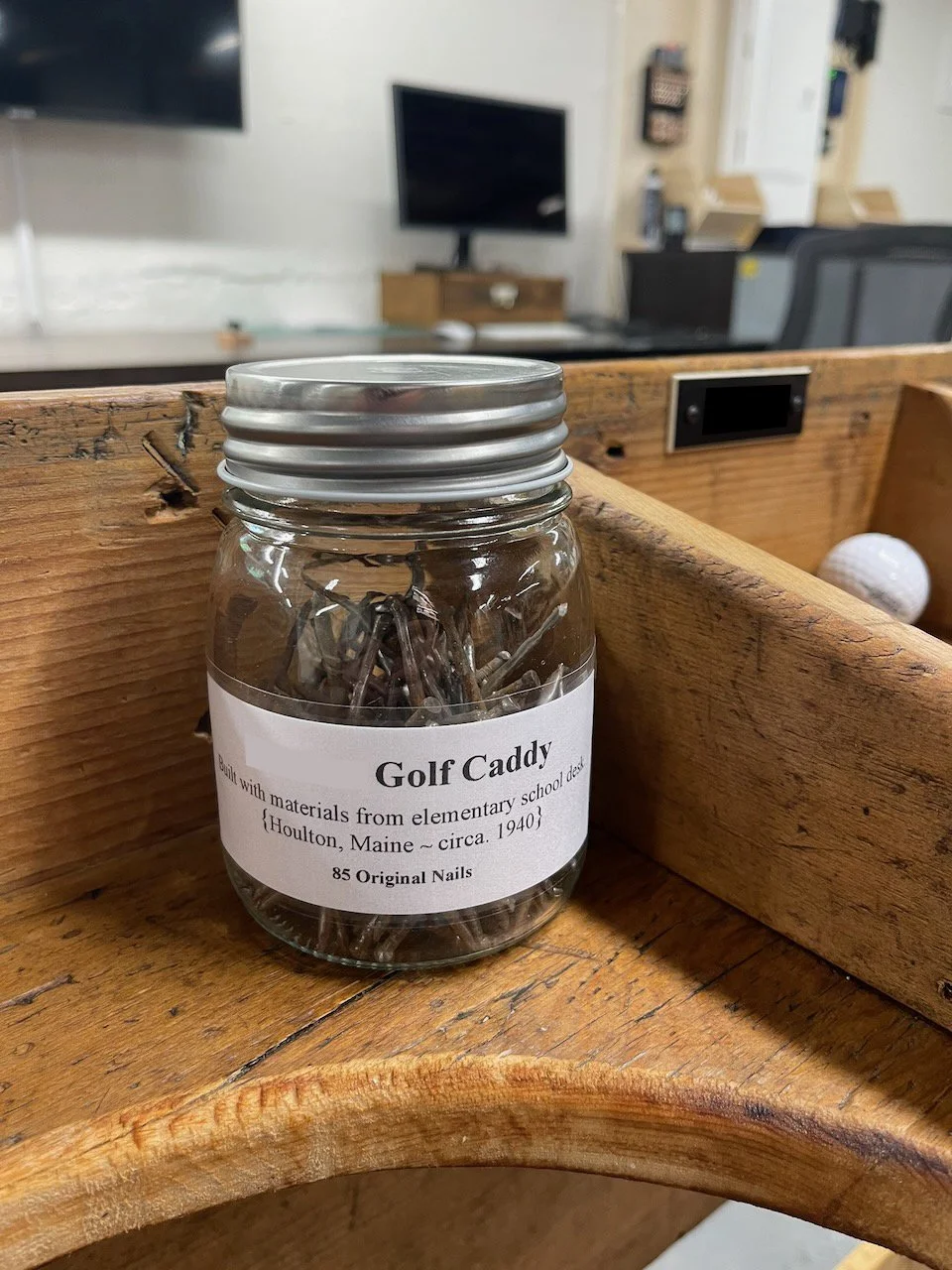 A glass jar filled with old nails, labeled 'Golf Caddy' and notes about materials from an elementary school desk in Houlton, Maine, circa 1940, on a wooden surface in a room with a TV and office furniture in the background.