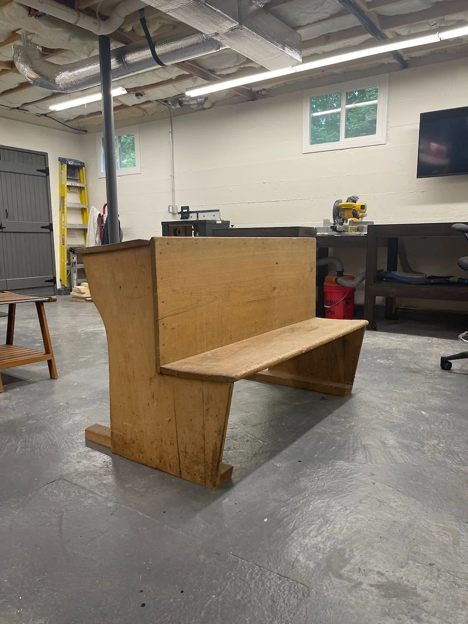 Unfinished wooden church pew in a workshop or garage with tools and equipment visible in the background.