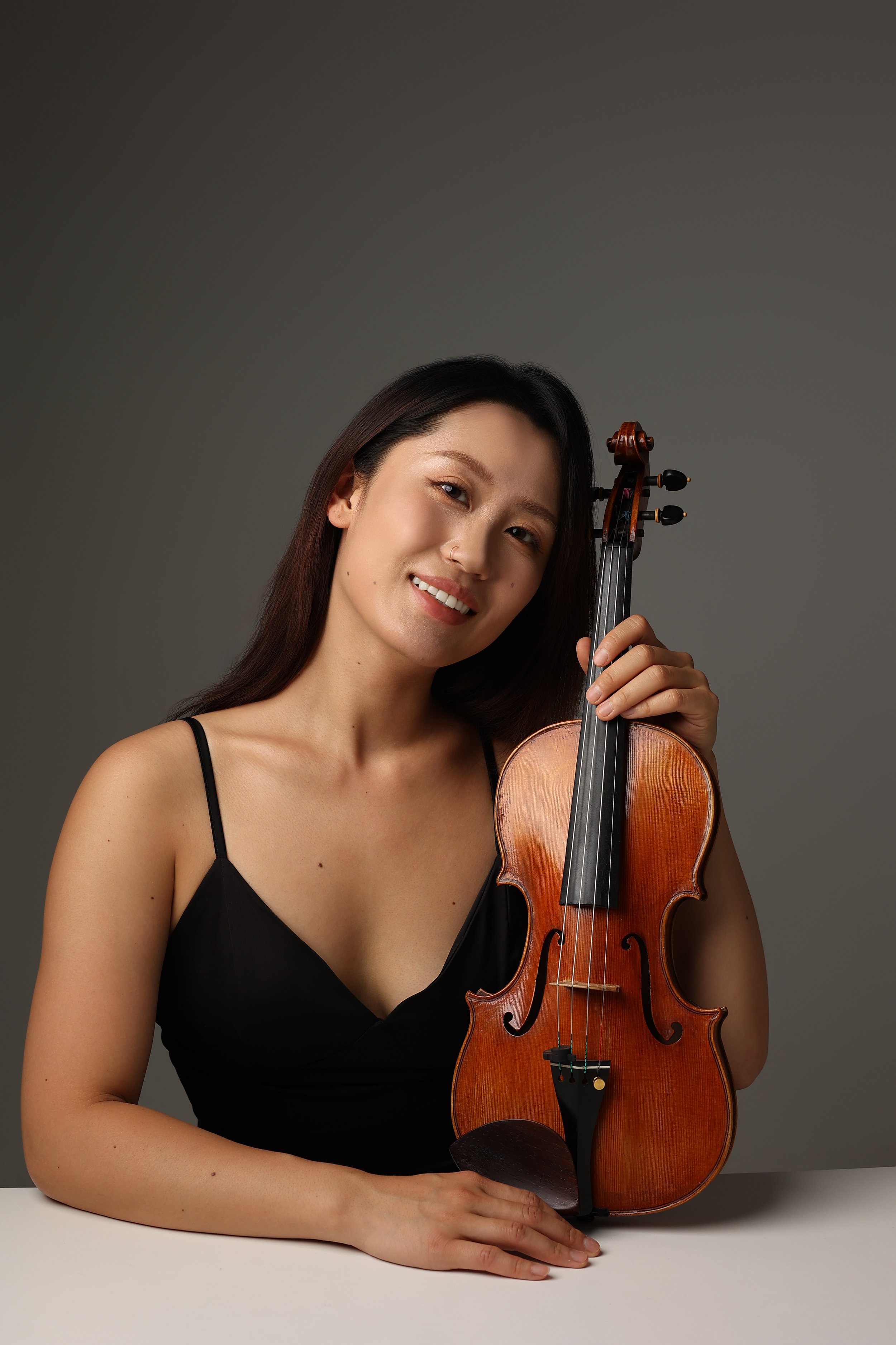 A woman with long dark hair in a black spaghetti strap top sits at a white table, holding a violin, smiling at the camera against a gray background.