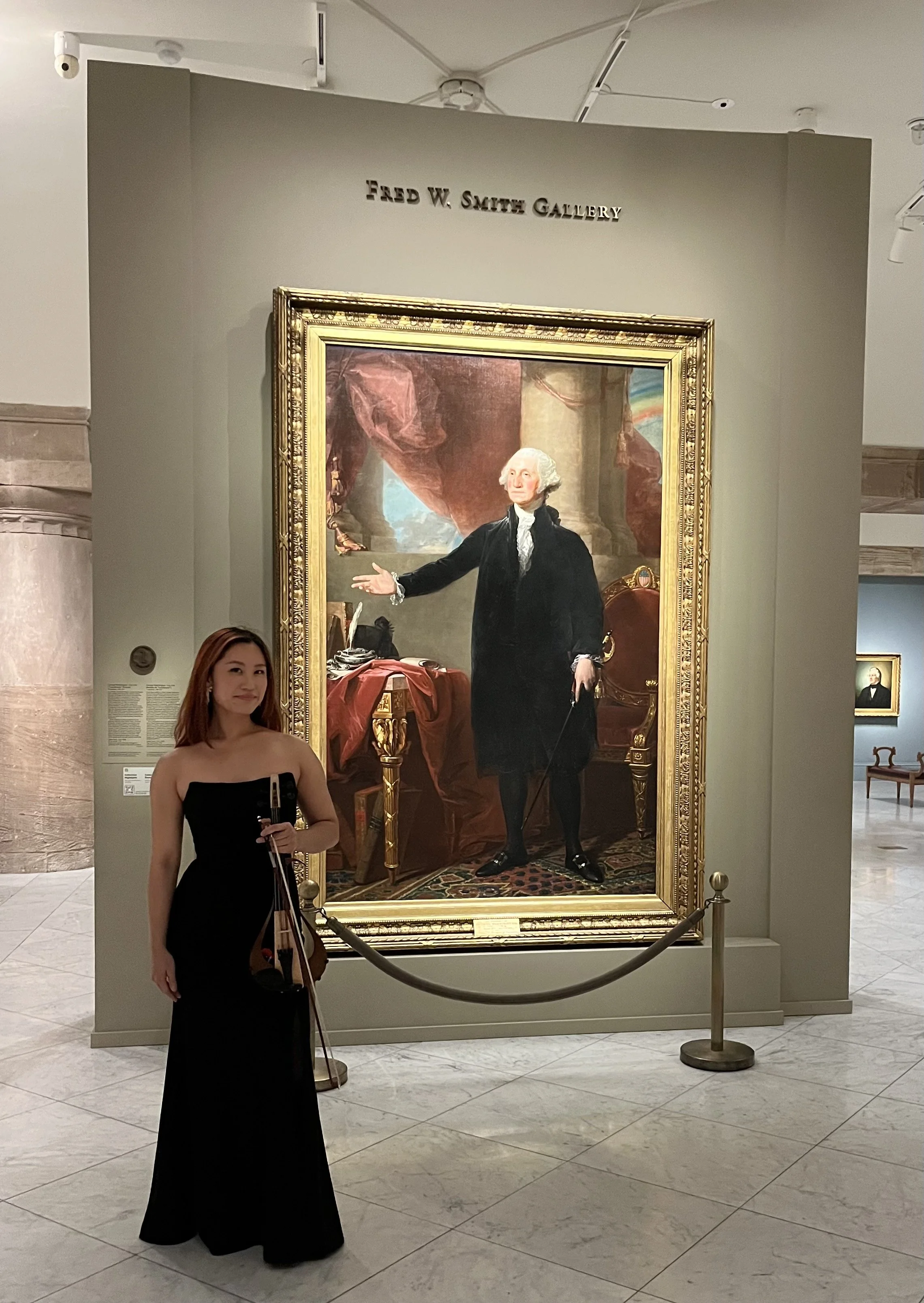 A young woman in a black strapless dress standing in front of a portrait of George Washington in the Fred W. Smith Gallery. The woman holds a violin, and the portrait of George Washington is displayed behind a barrier in an art museum.