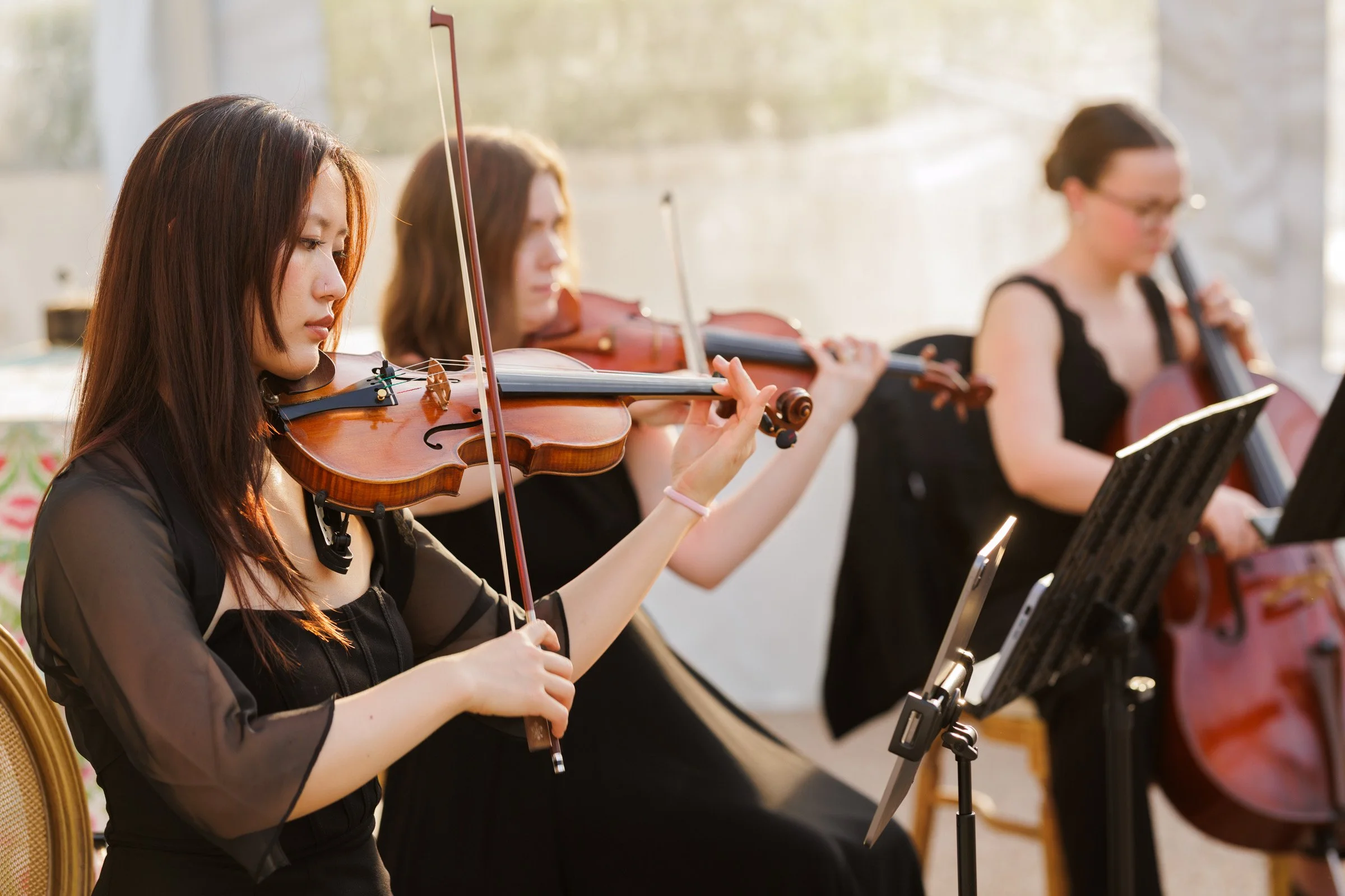 Women playing violins in a concert, sitting on chairs with music stands and reading music.