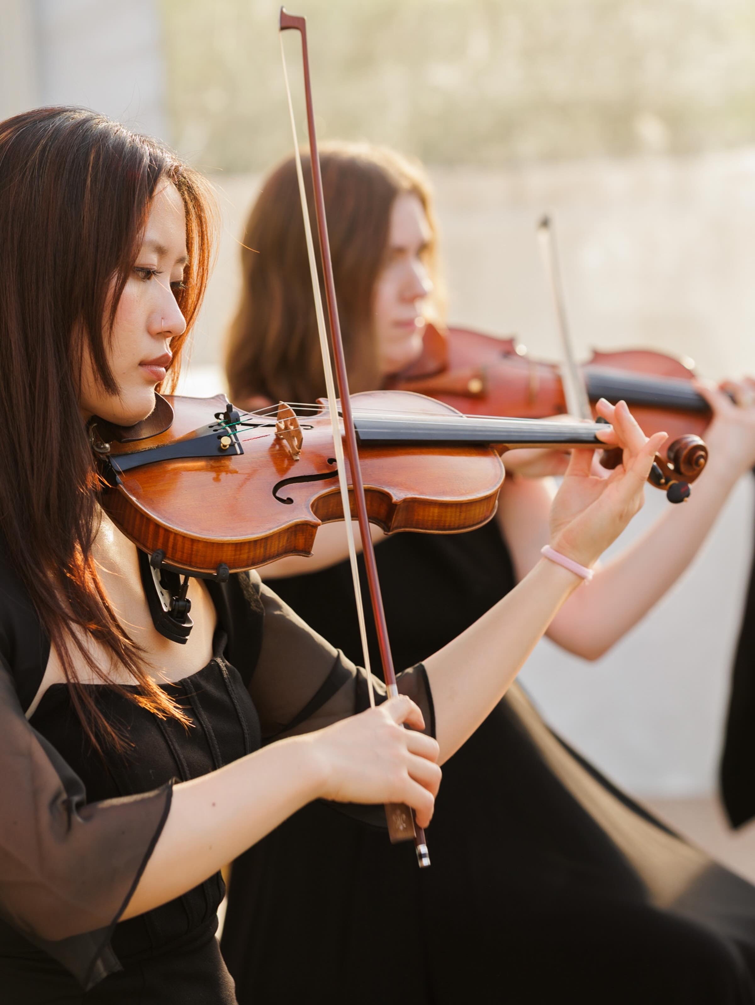 Two women playing the violin outdoors in sunlight, focusing on their bows and violins.