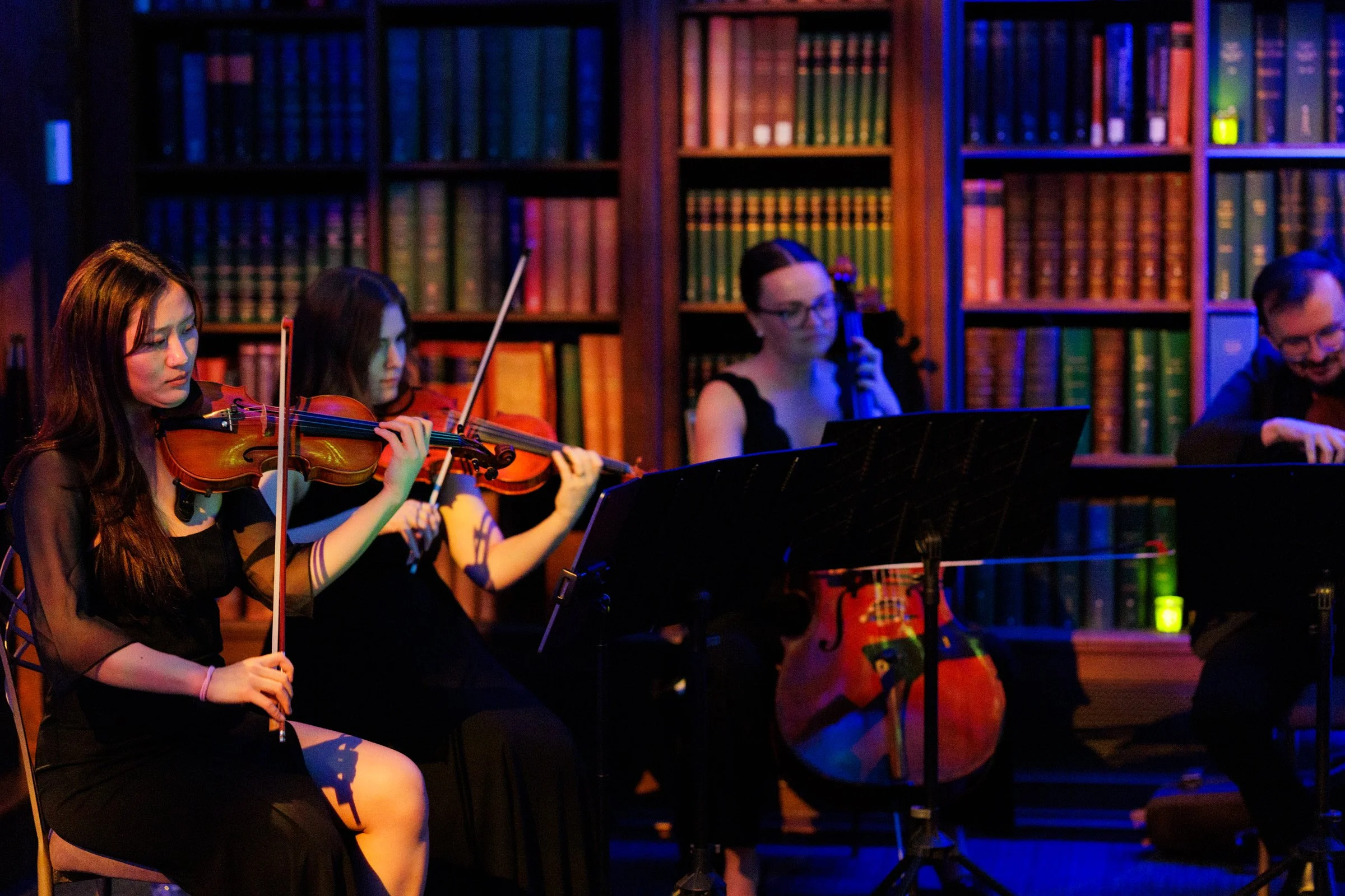 Young women playing violins in a dimly lit room with bookshelves in the background.