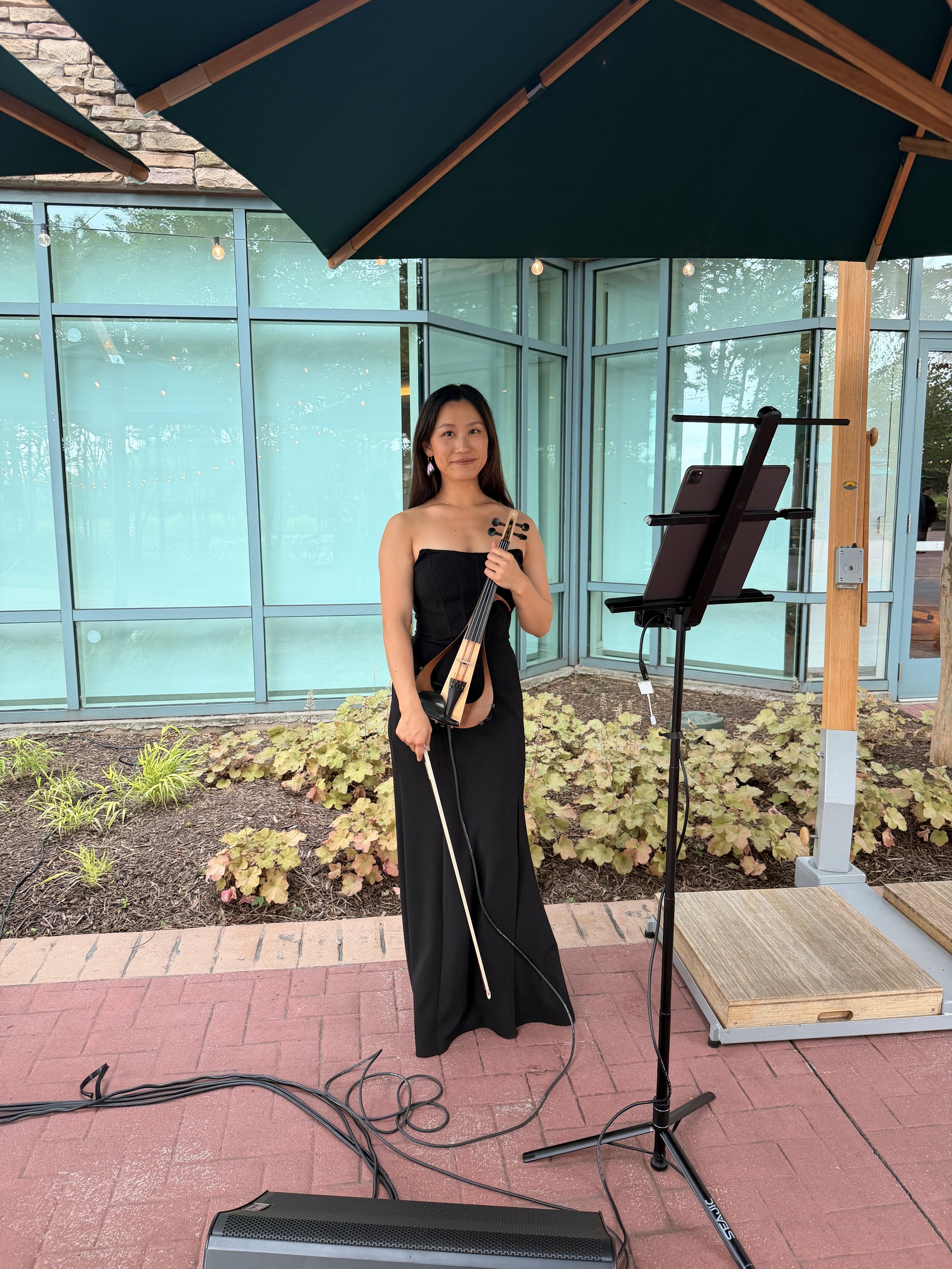 A woman in a strapless black dress holding a cello and bow, standing under a large green patio umbrella outside a glass building with string lights, plants, and a brick and sidewalk ground.