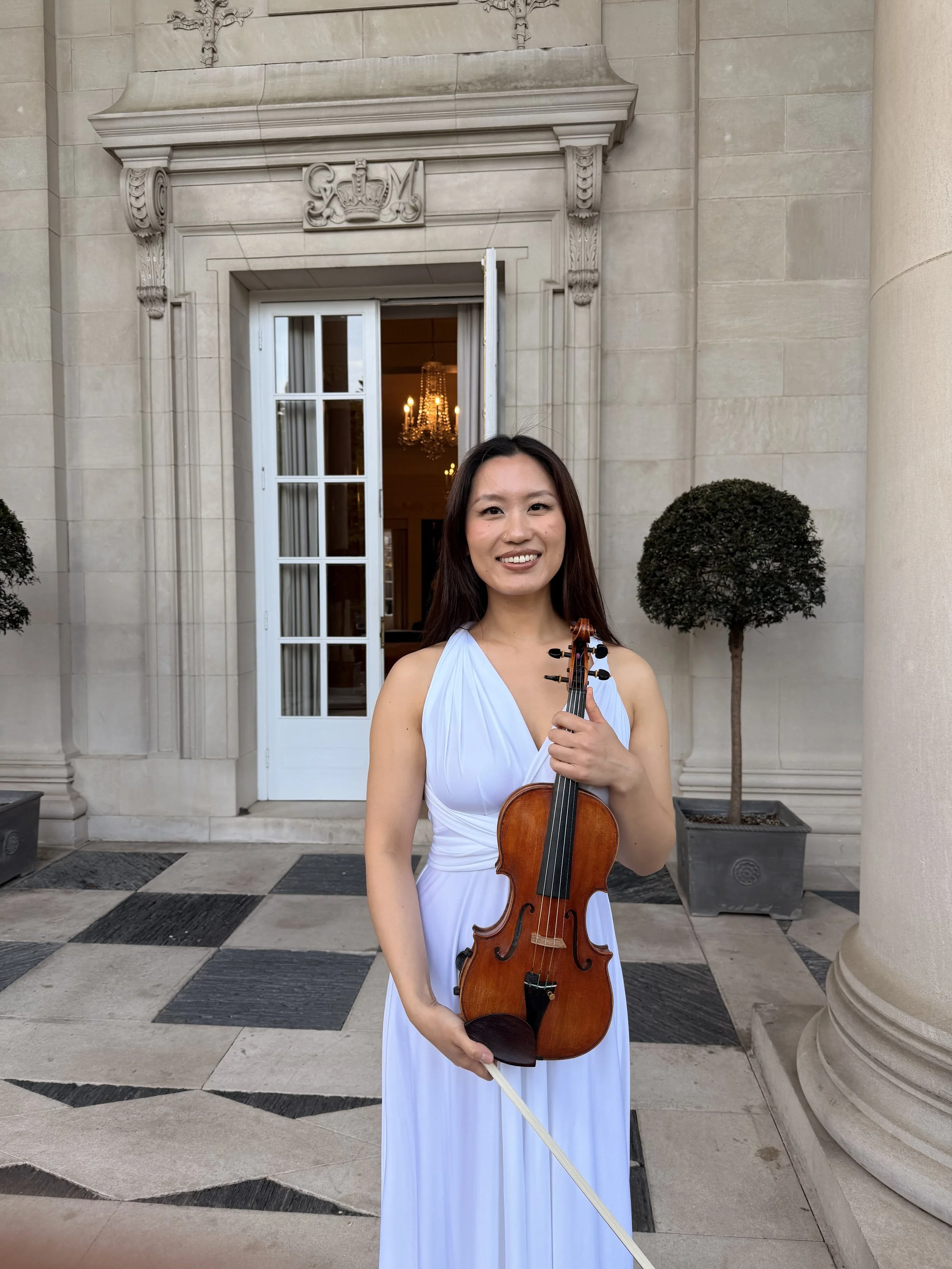 A woman in a white dress holding a violin and bow, standing outside a building with stone walls, a glass door, and potted trees.