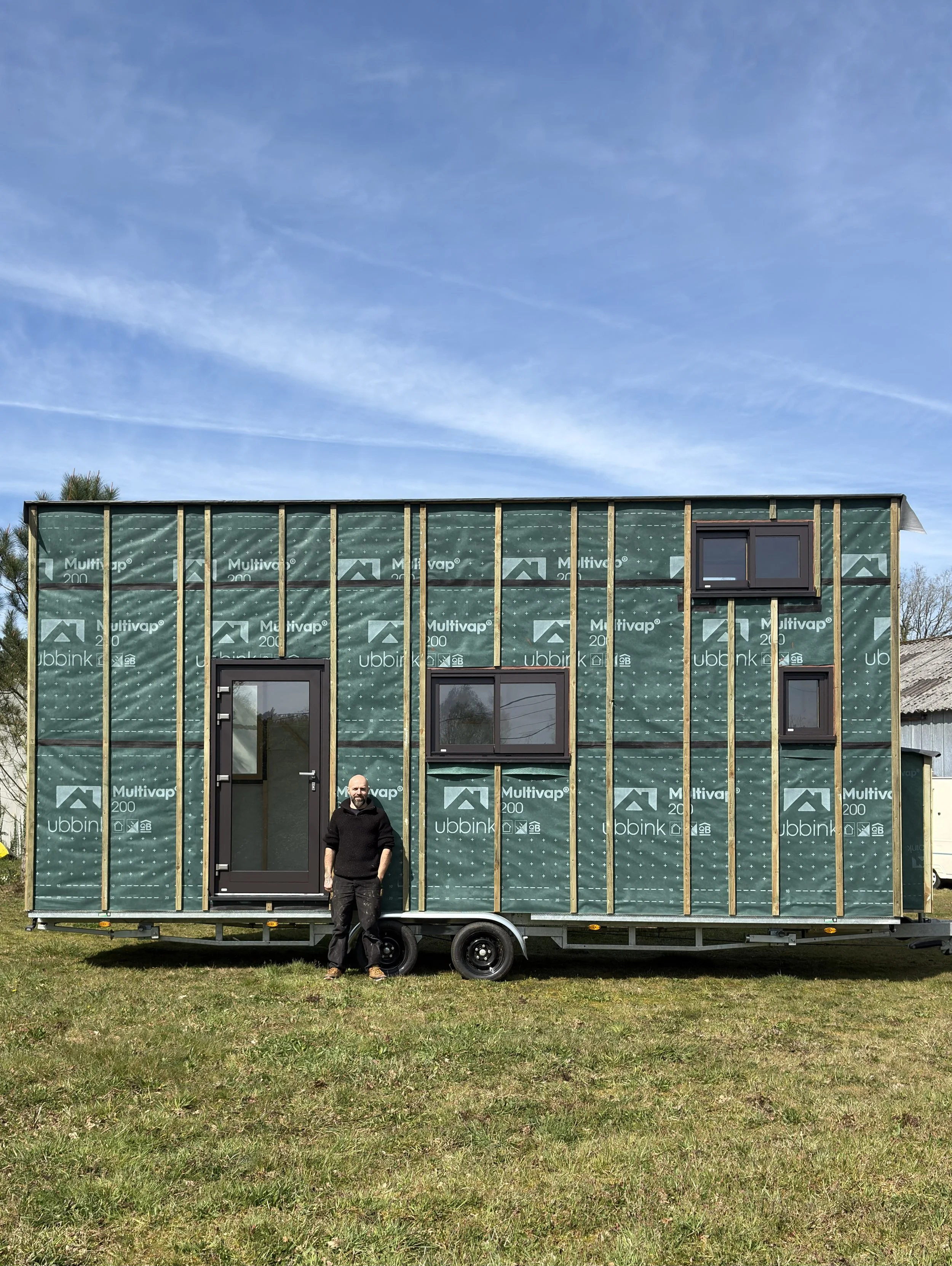 Une maison en construction sur une remorque. La maison a une structure en bois et est recouverte d'un pare-pluie. Il y a plusieurs fenêtres mixte bois aluminium installées.
