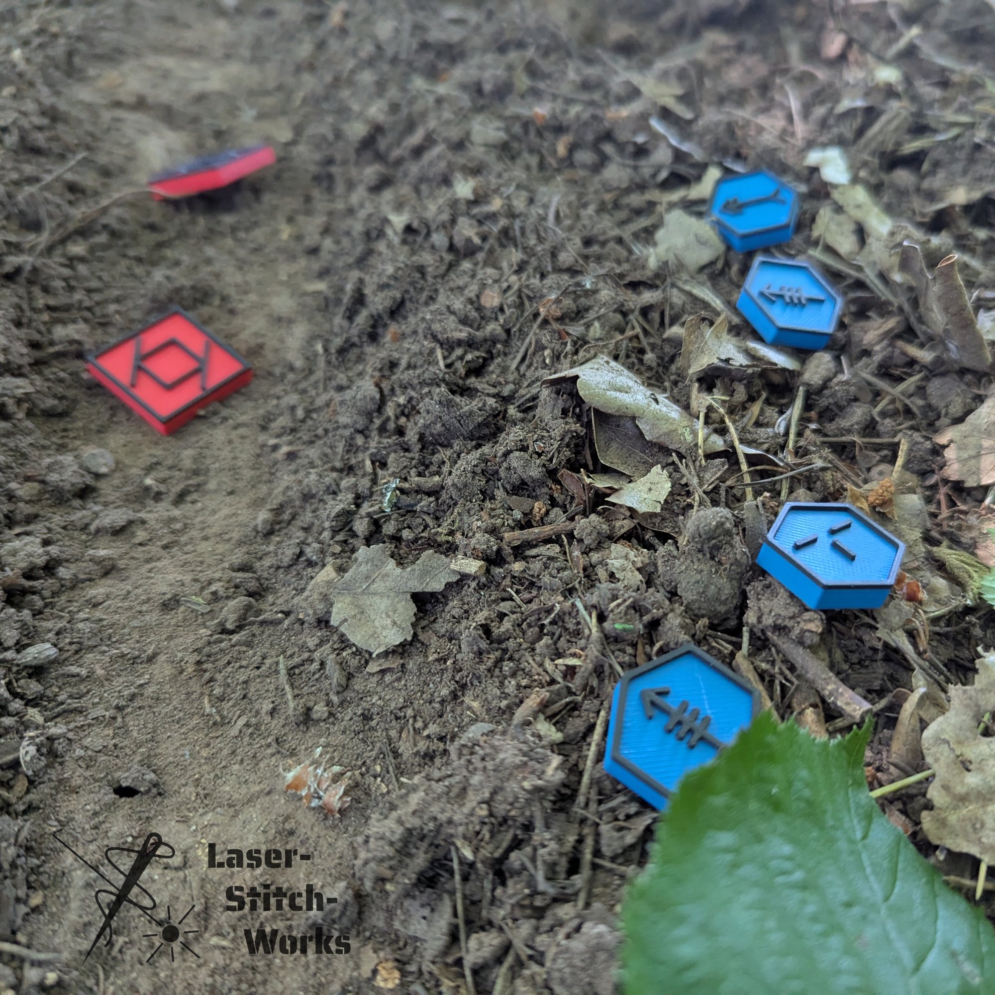 Boden mit Erde, Blättern und kleinen Plastikteilen in Form von Spielkacheln, die Zahlen und Symbole darstellen, in blau und rot.