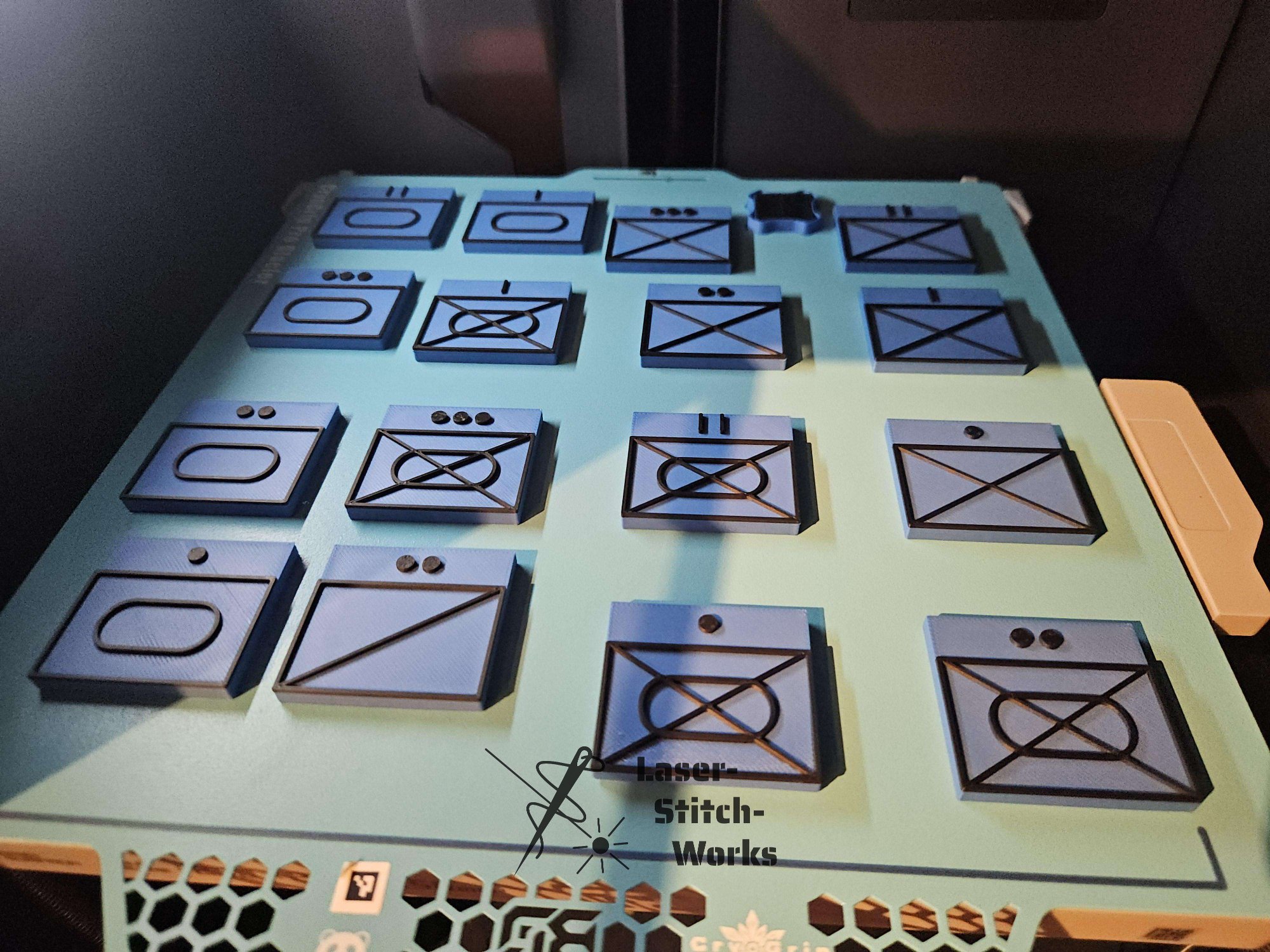 Schwarzes Brettspiel mit quadratischen Spielfeldern, das auf einer grünen Oberfläche liegt. Die Felder sind mit Linien und Symbolen beschrieben, einige sind ausgelegt für spezielle Spielzüge. Es gibt eine Marke von Laser Stitch Works auf dem Brett.